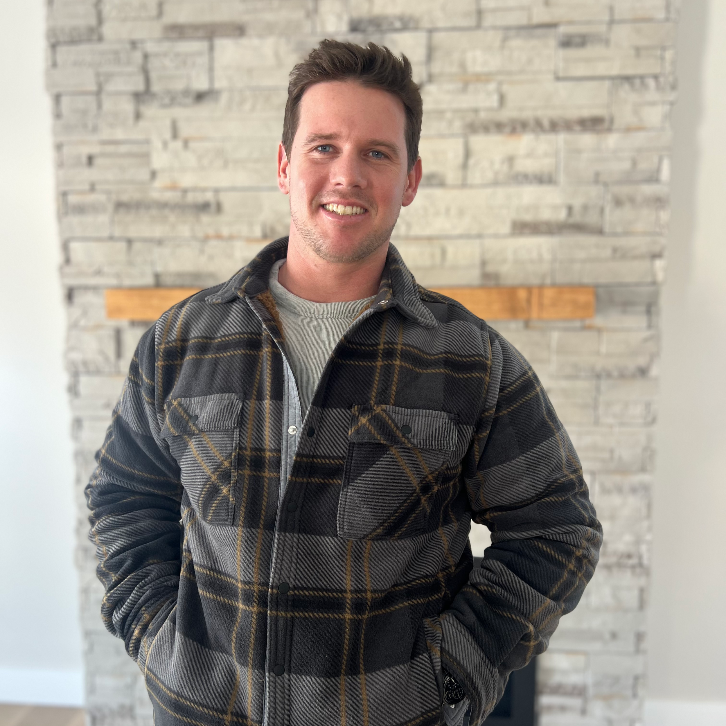 A young man with short brown hair smiling, wearing a plaid jacket over a light grey shirt, with a white brick fireplace in the background.
