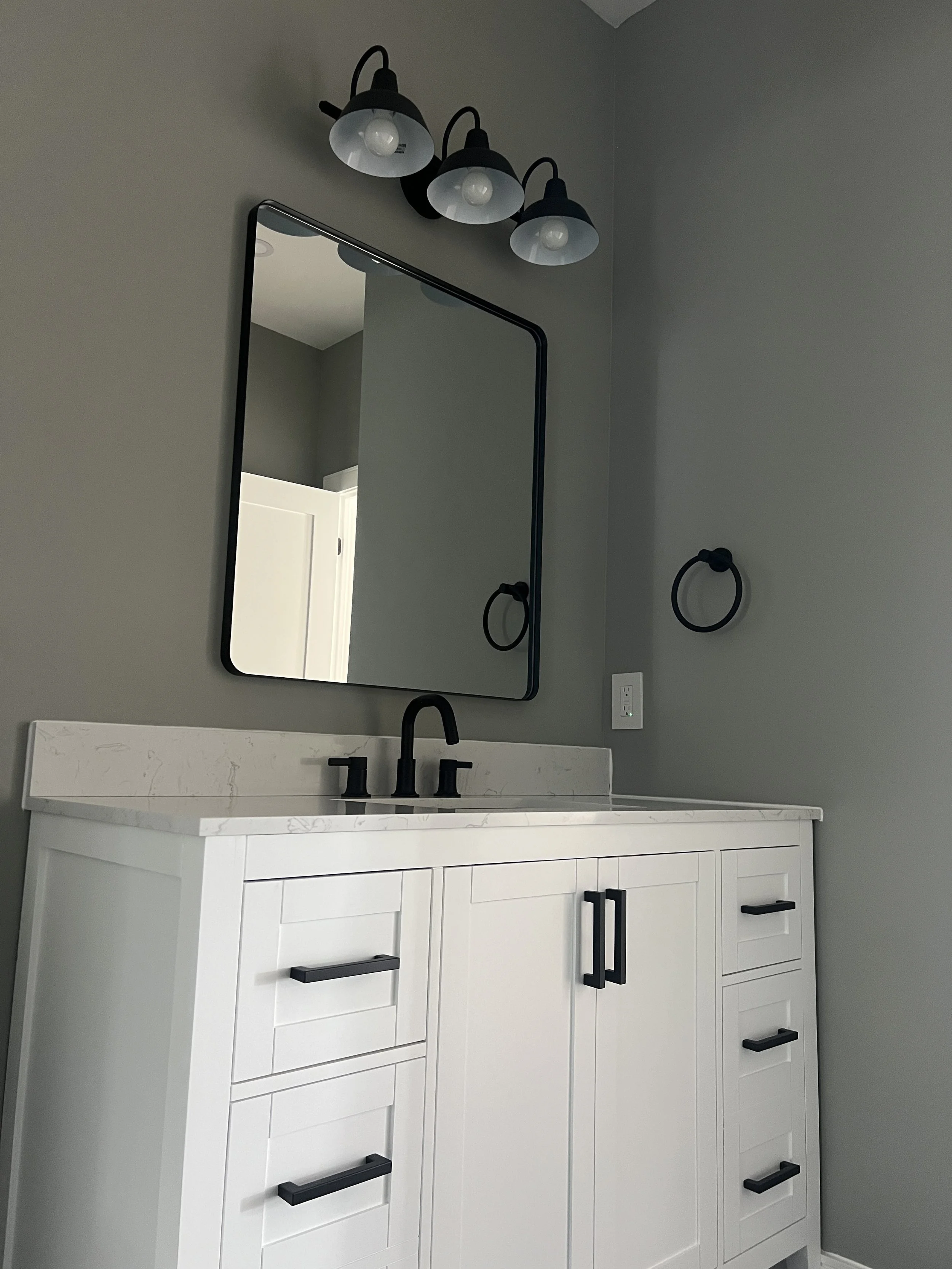 Modern bathroom vanity with a white cabinet, black handles, a marble countertop, a black faucet, a rectangular mirror, and a black three-light fixture above the mirror.
