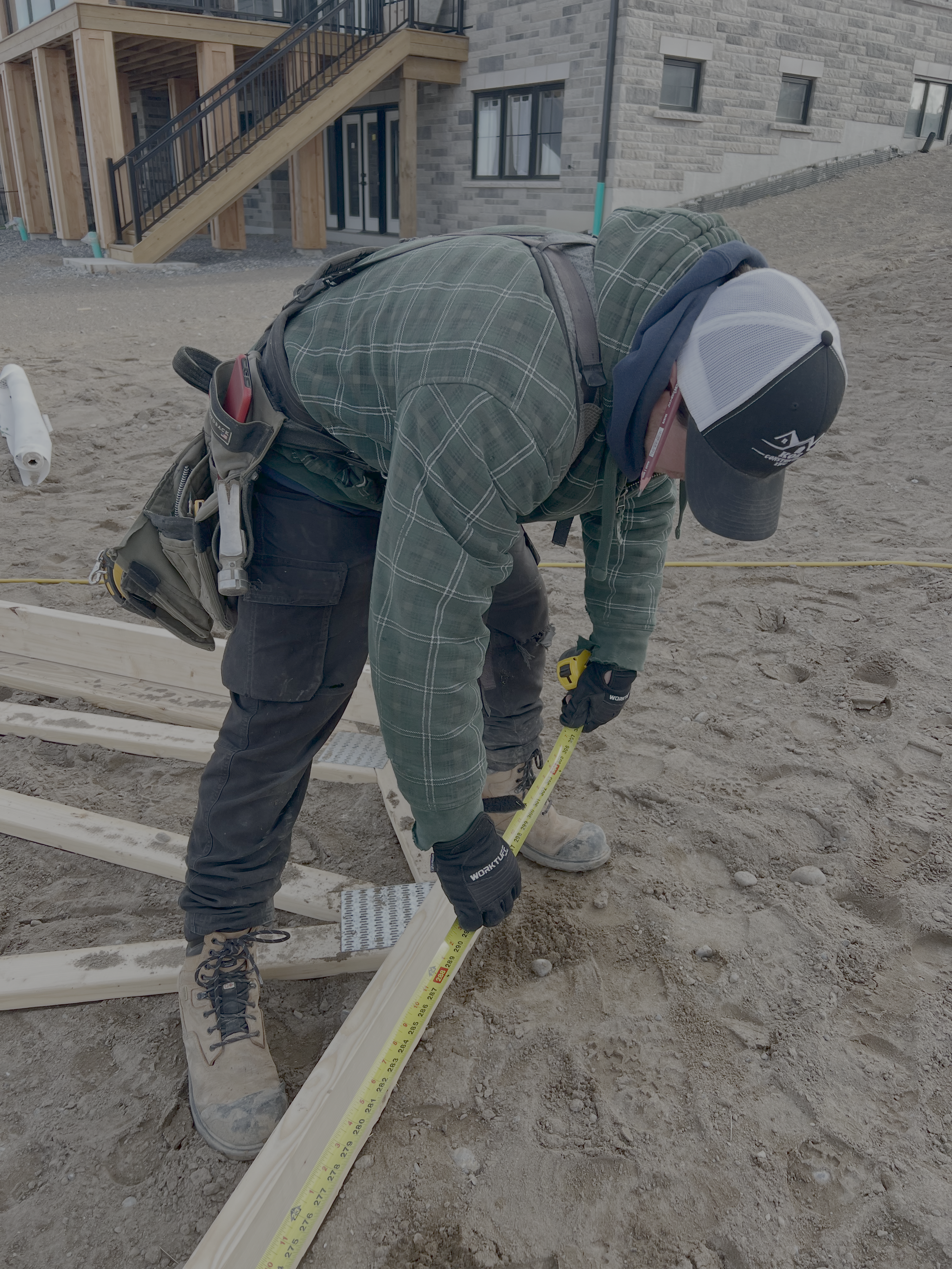 Construction worker measuring wooden board on uneven dirt ground outside a building under construction.