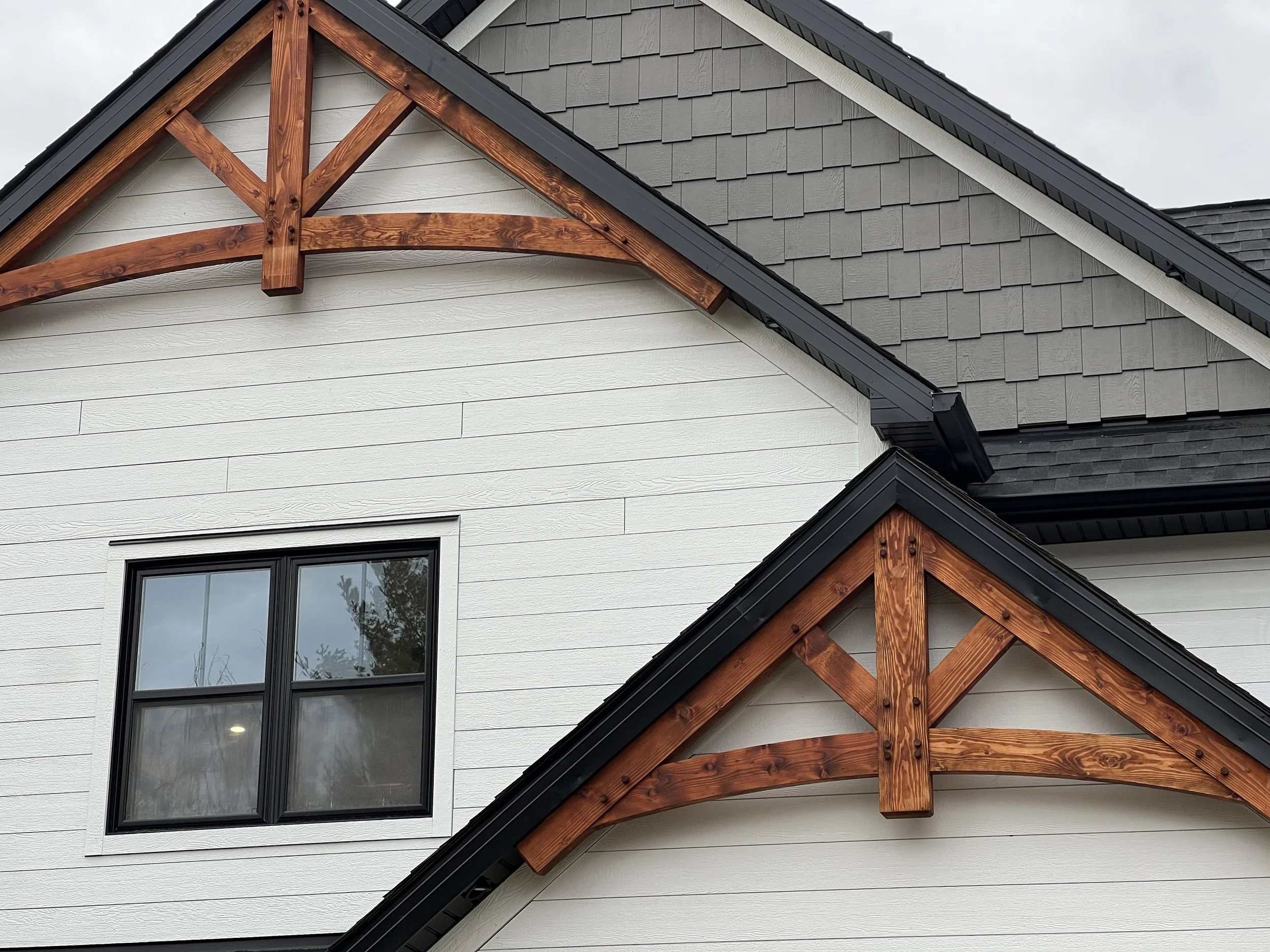 Close-up of the exterior of a modern farmhouse showing white siding, black window frame, and decorative wooden gables with stain finish, along with black roof shingles and black trim.