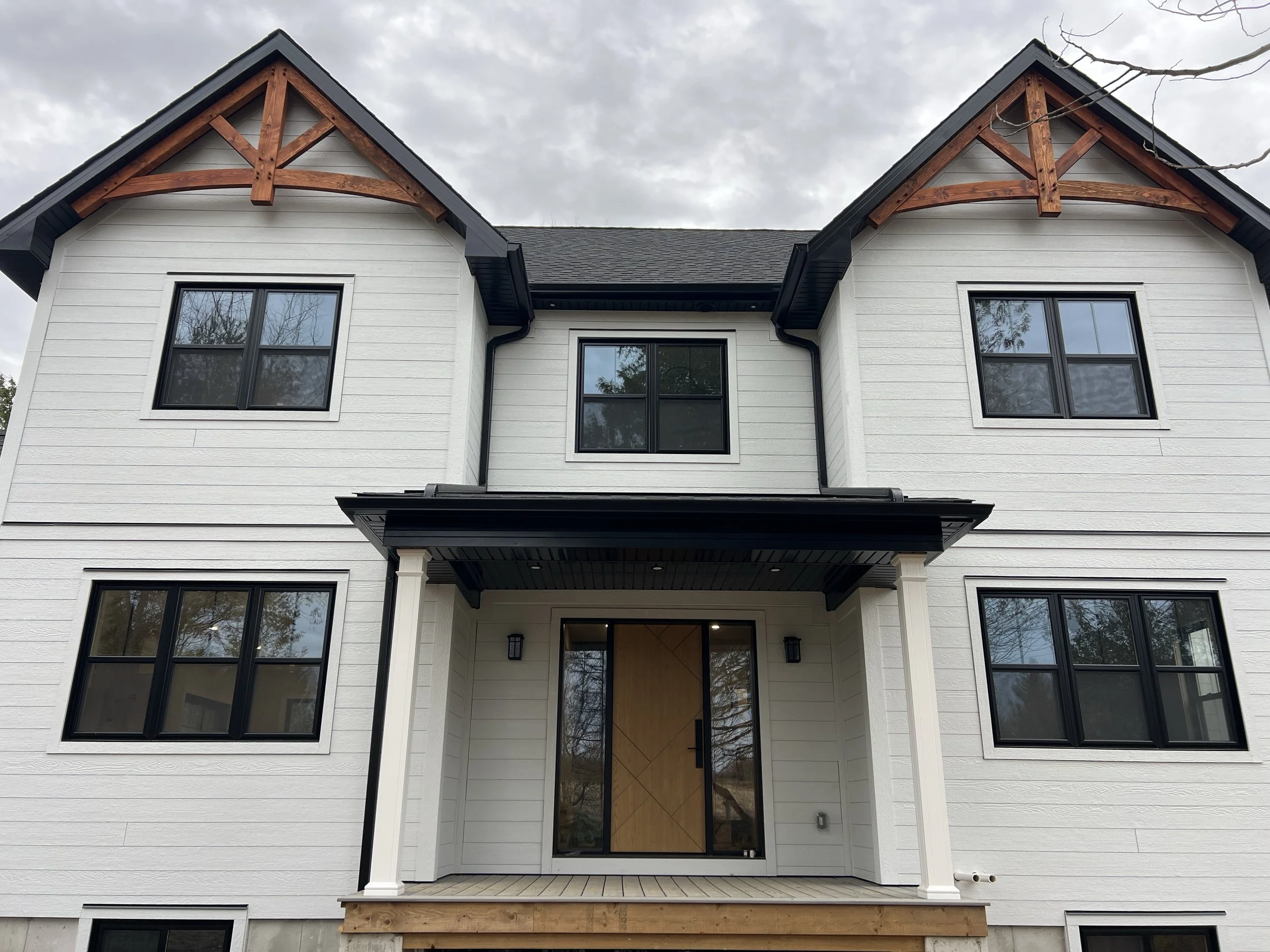 Newly built white two-story modern farmhouse with black window frames, a front porch with columns, and decorative wooden gables with stain finish.
