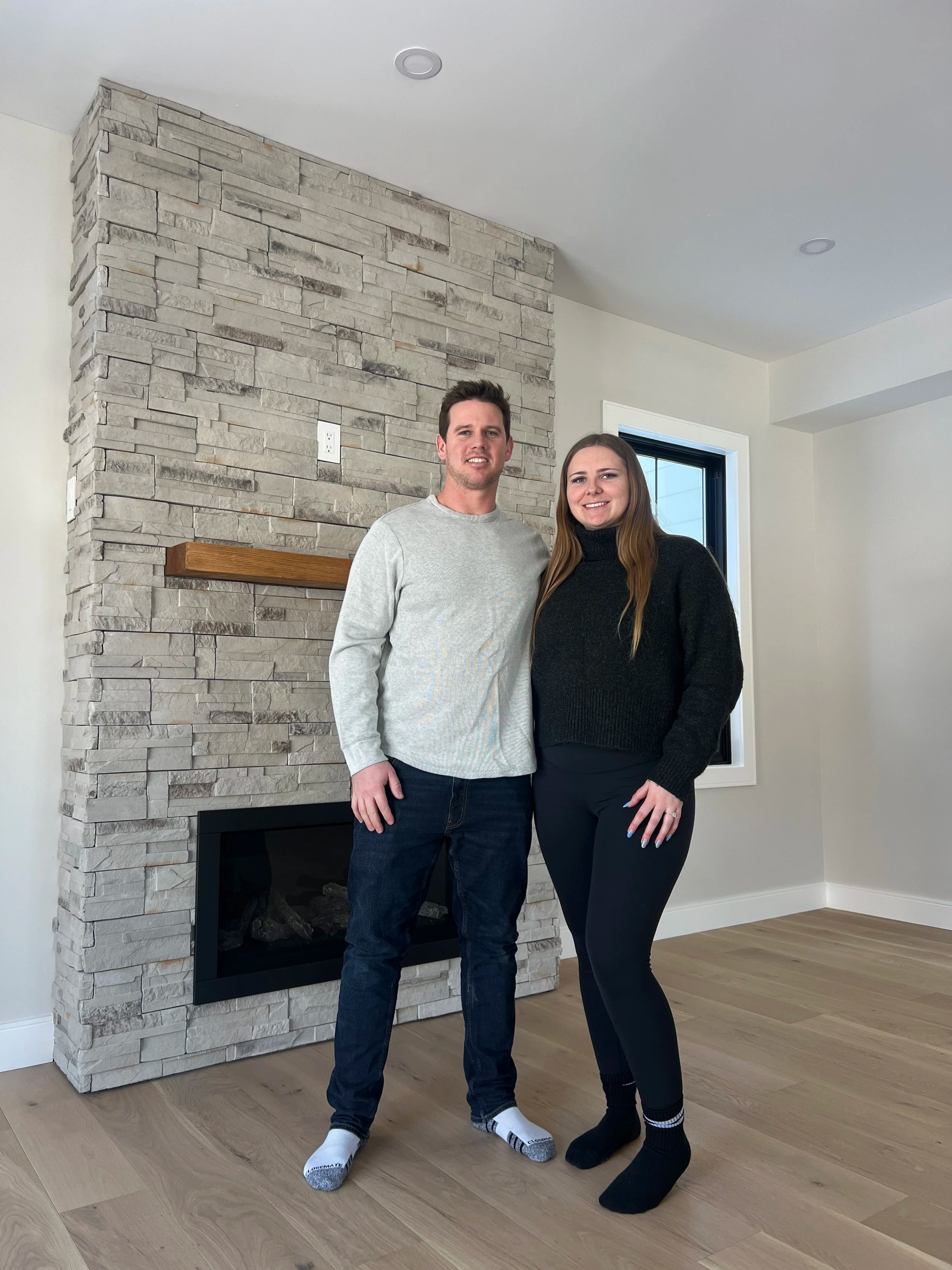 A man and woman standing side by side indoors, in front of a stacked stone fireplace with a wooden mantel, a window with a black frame, and a light-coloured wall.