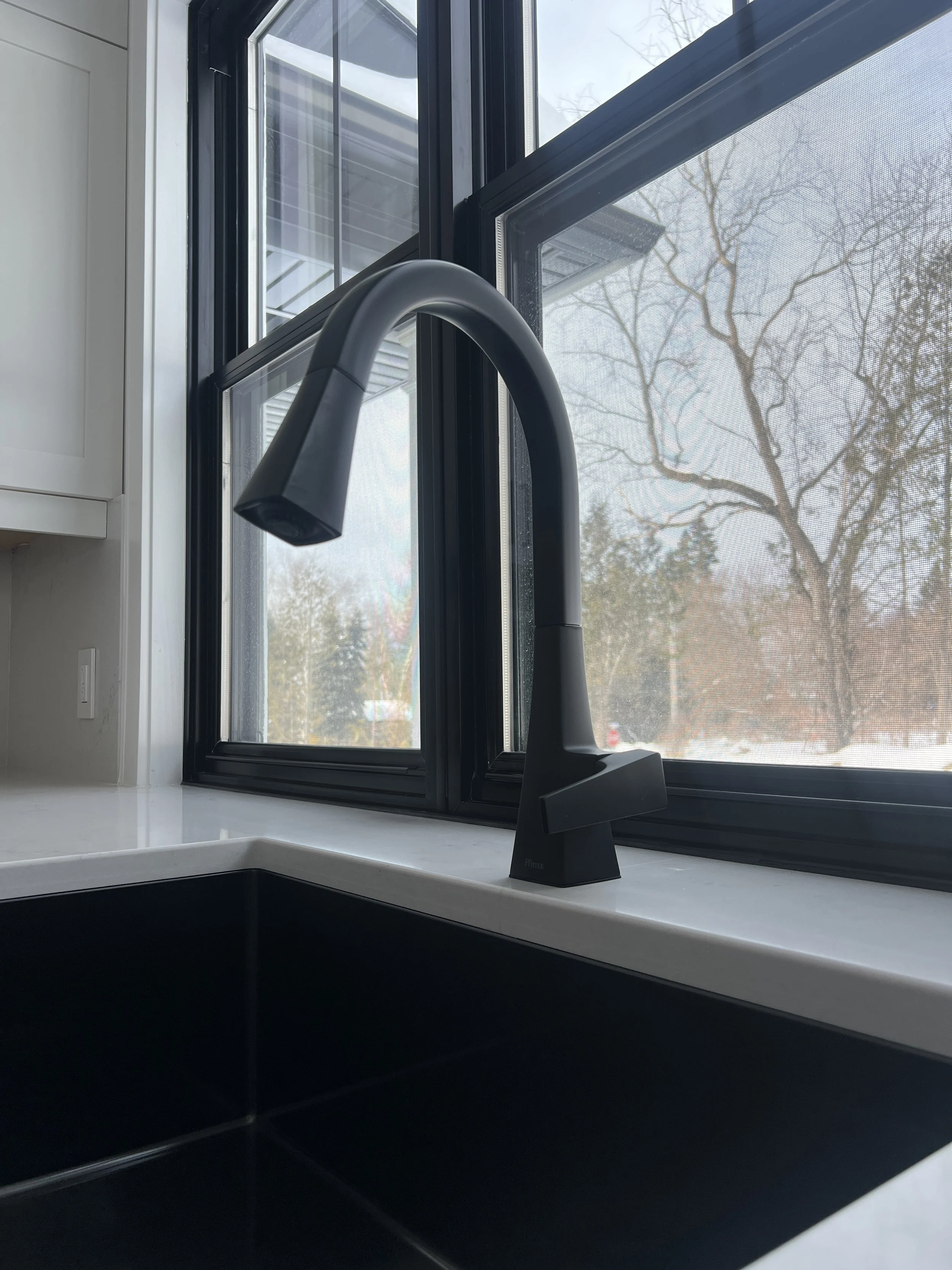 Close-up of a black kitchen faucet on a white countertop, with a large black-framed window.