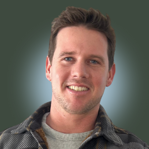 A smiling man with light brown hair, wearing a wearing a plaid jacket against a green background.