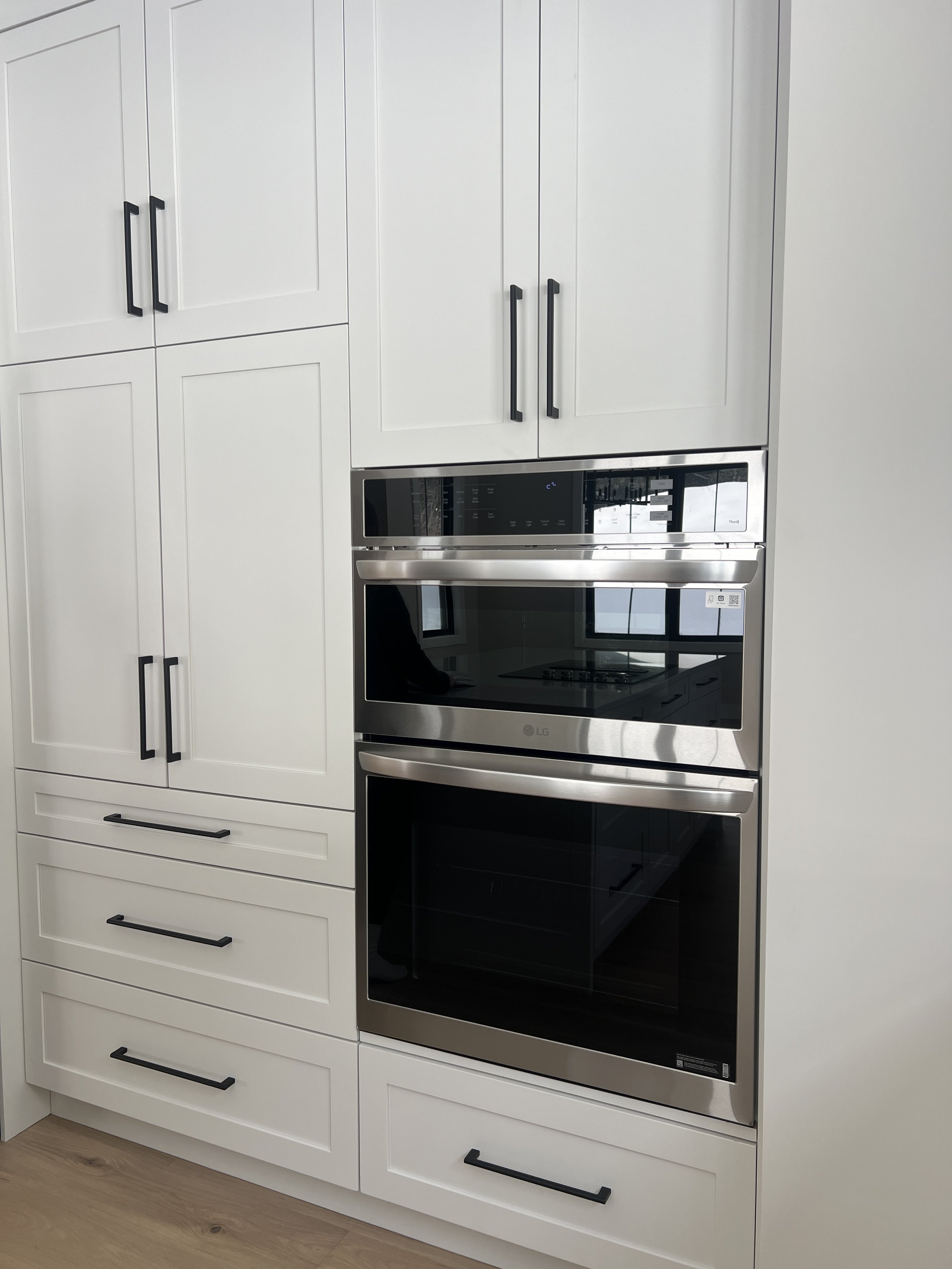 Built-in wall oven and microwave in a modern kitchen with white cabinets and black handles.