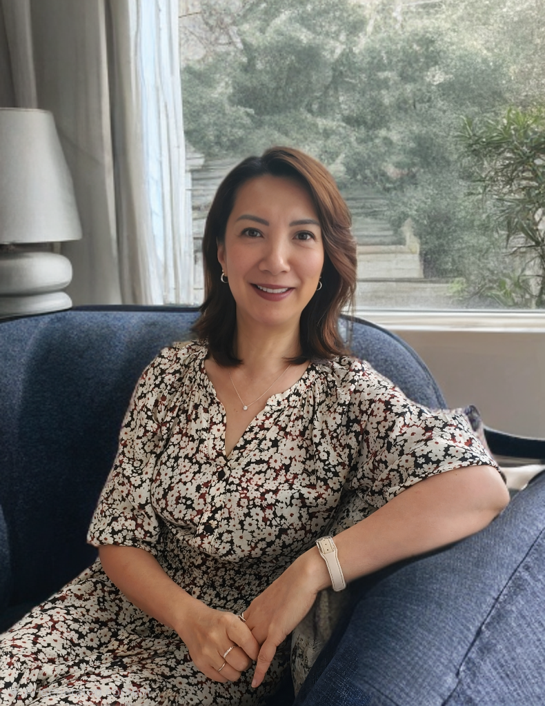A woman sitting on a blue armchair in front of a window, smiling, with a white lamp and greenery outside.