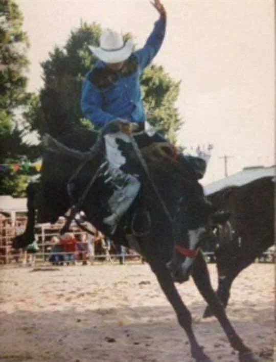 Person wearing a cowboy hat and blue shirt riding a bucking horse at a rodeo.