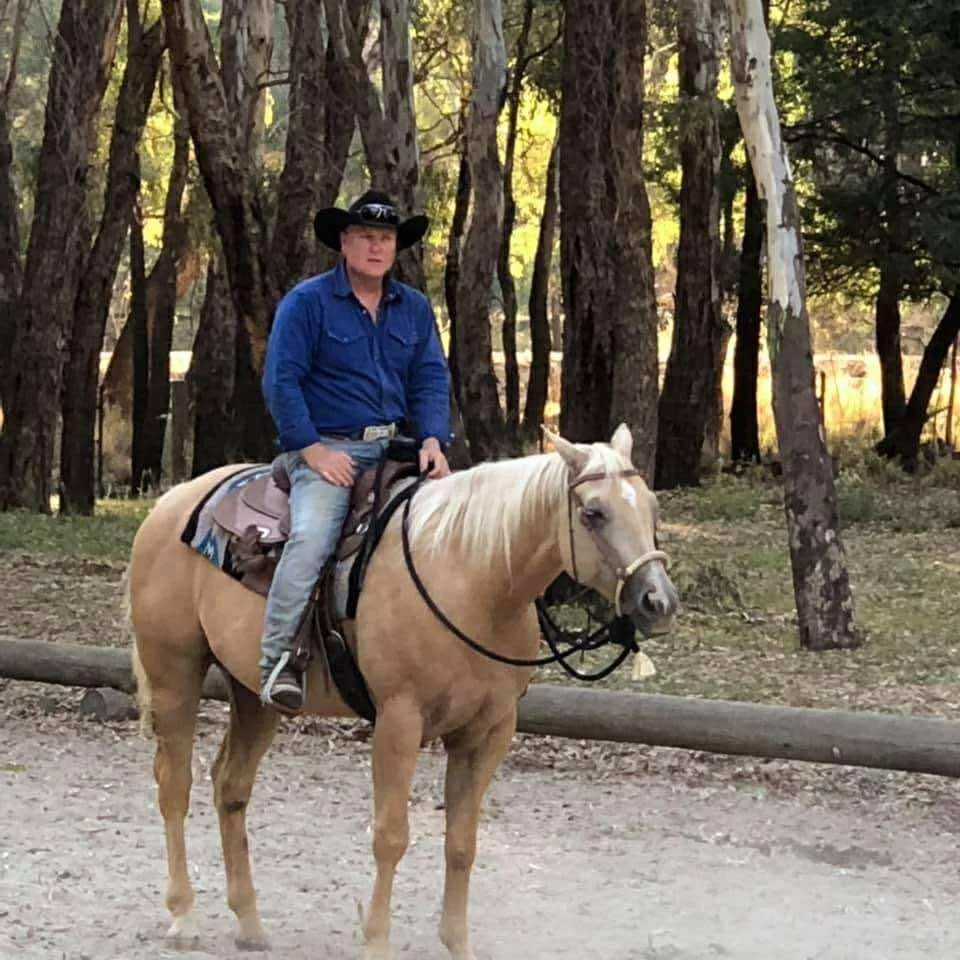 A person wearing a cowboy hat and blue shirt riding a light-colored horse in a wooded area.