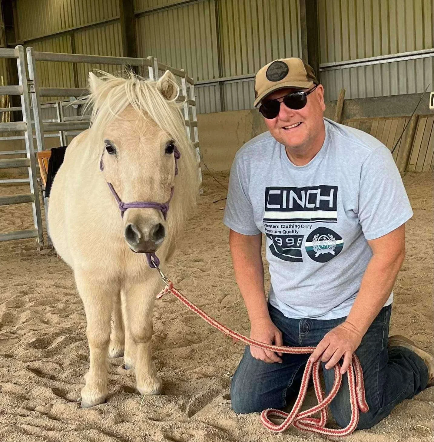 A man kneeling on the ground with a white horse indoors in a riding arena, smiling and holding the horse's lead rope.