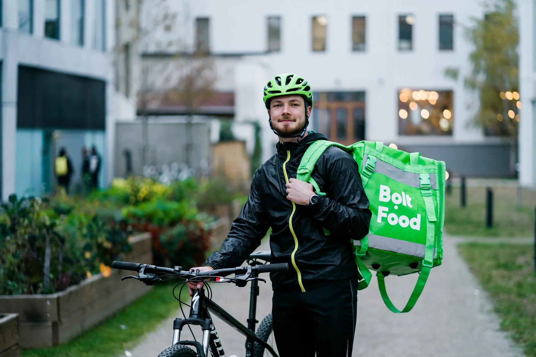 A man with a beard in a yellow-green helmet and black jacket standing with a mountain bike, carrying a green Bolt Food delivery backpack, in an urban park setting. Advertising campaign photographer for corporate and startup business Switzerland.