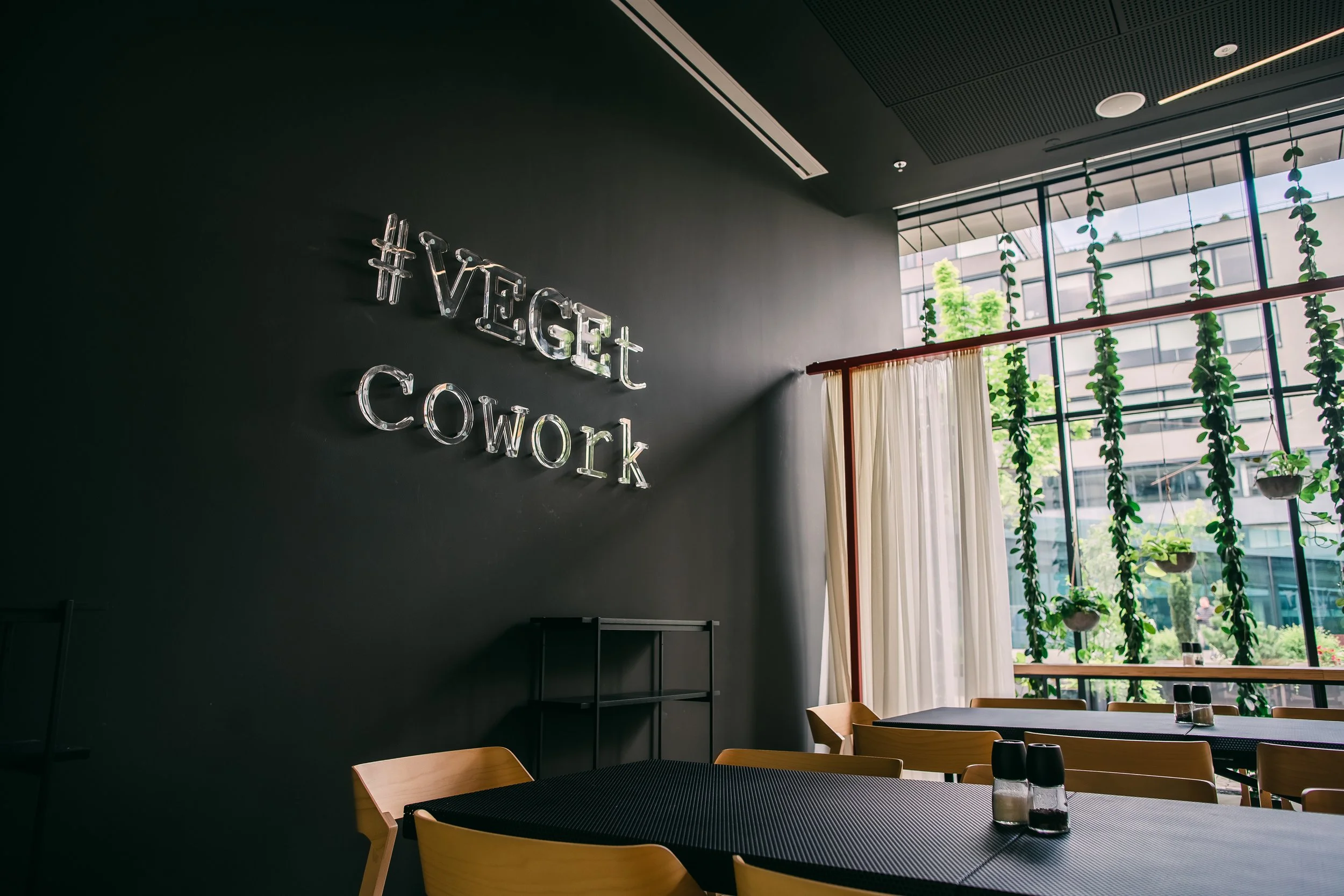 Modern dining area with wooden chairs and tables, neon sign on dark wall reading '#VGET COWORK,' large windows with curtain and hanging plants, natural light, urban city view. Restaurant interior photography in Zürich, Zug, Switzerland.