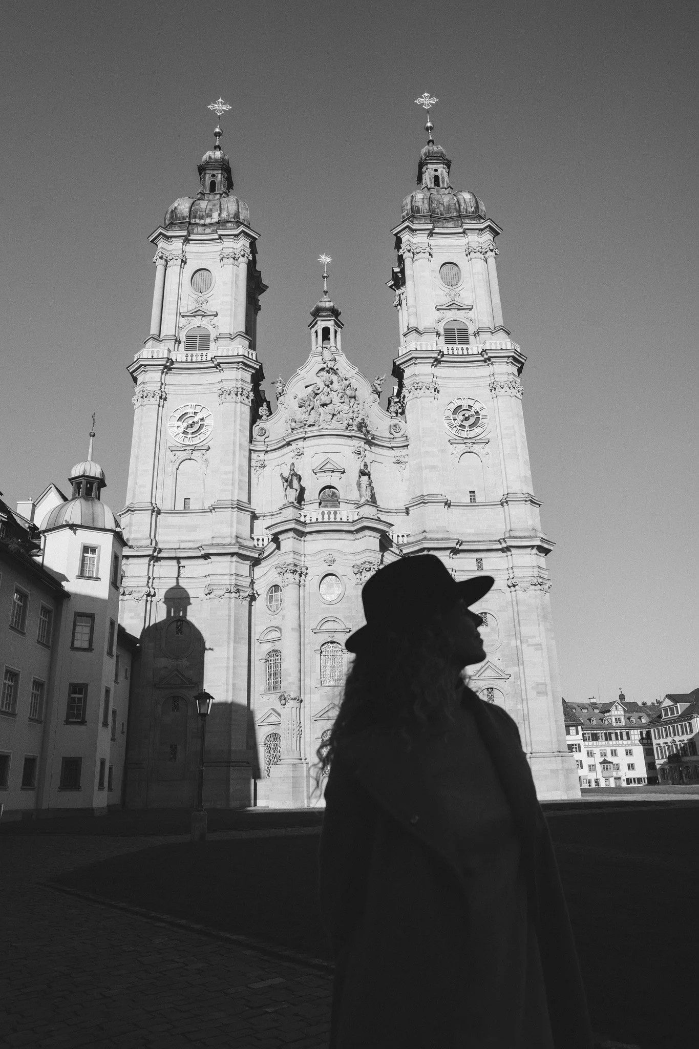 Silhouette of a woman wearing a hat in front of a church with twin towers and Baroque architecture, with a clear sky background. Artsy photography based in St. Gallen Switzerland.