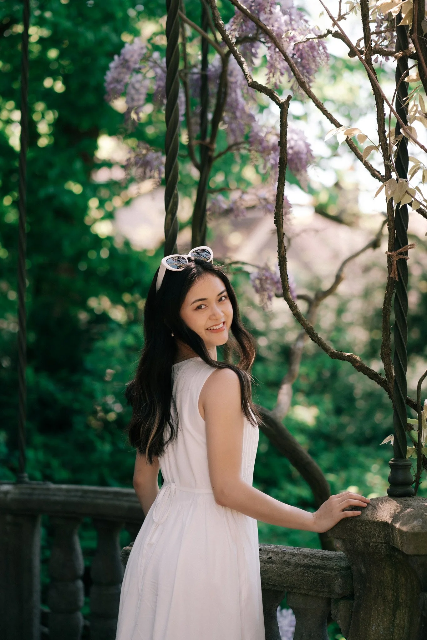 A woman in a white romantic dress standing in a garden in Zurich Switzerland with purple flowers and green foliage, smiling at the camera.