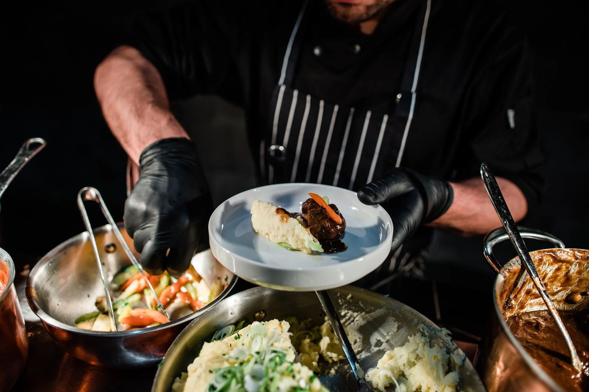 Chef in black clothing and gloves serving plated food at a buffet. Catering and food photography in Zürich, Zug, Bern, Lucerne, Basel, St. Gallen and across Switzerland.