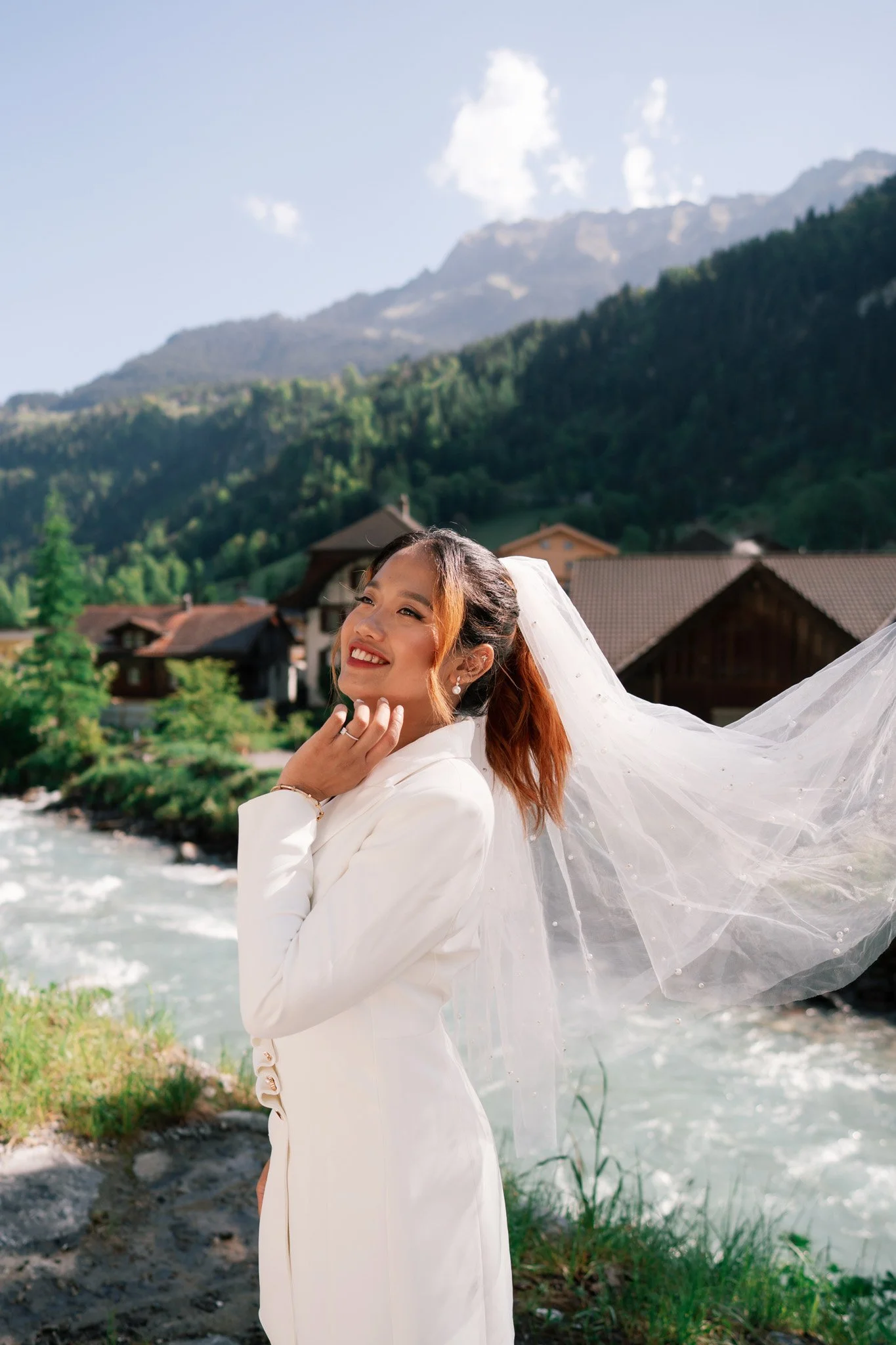 A woman in a white wedding dress and veil smiling outdoors for engagement photoshoot in Lauterbunnen, Switzerland, near a river with mountains in the background.