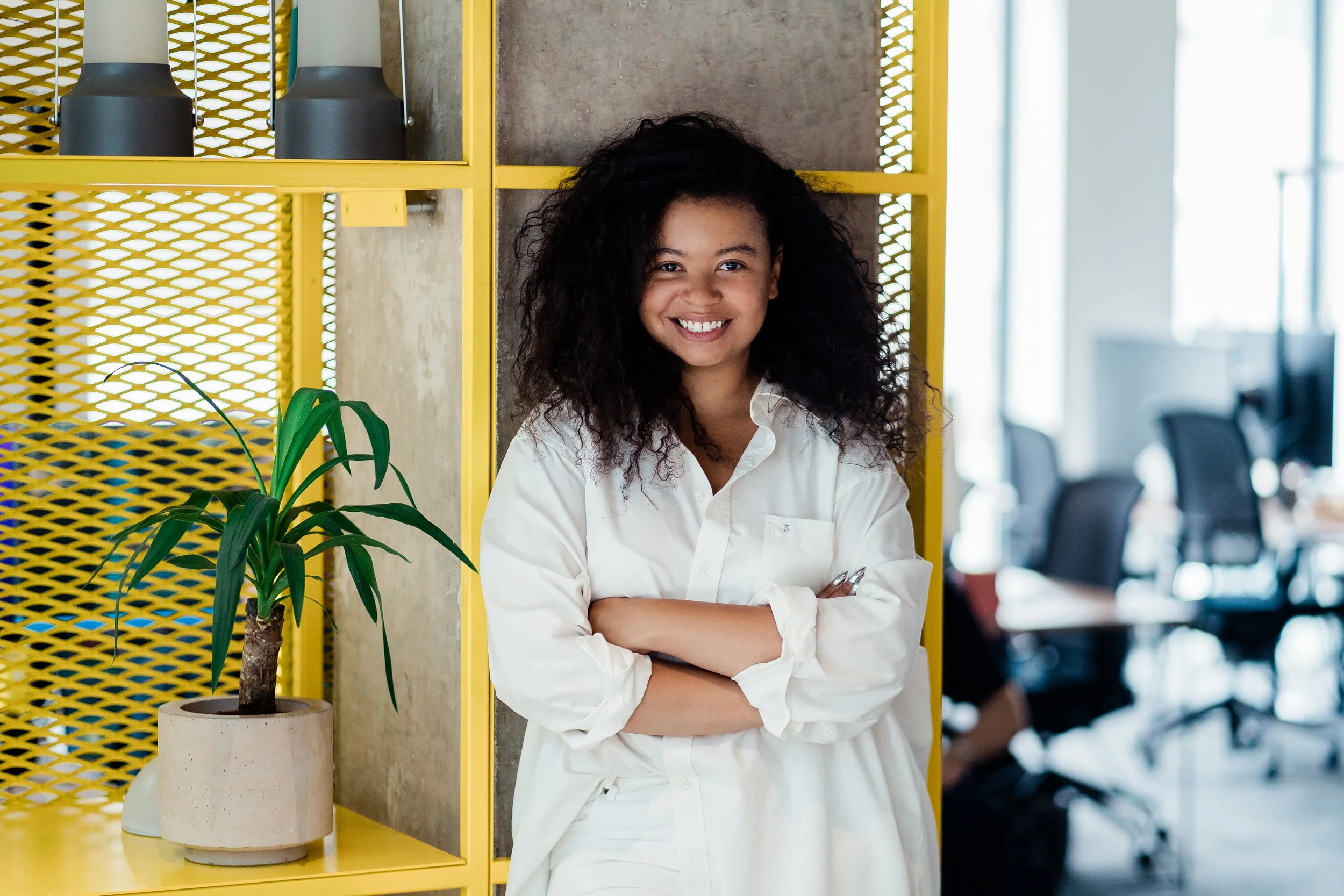 A woman with curly hair smiling and standing with arms crossed in a modern office space, yellow shelving, and a potted plant in the foreground. Corporate headshots photography in Zürich, Zug, Bern, Lucerne, Basel, St. Gallen and across Switzerland.