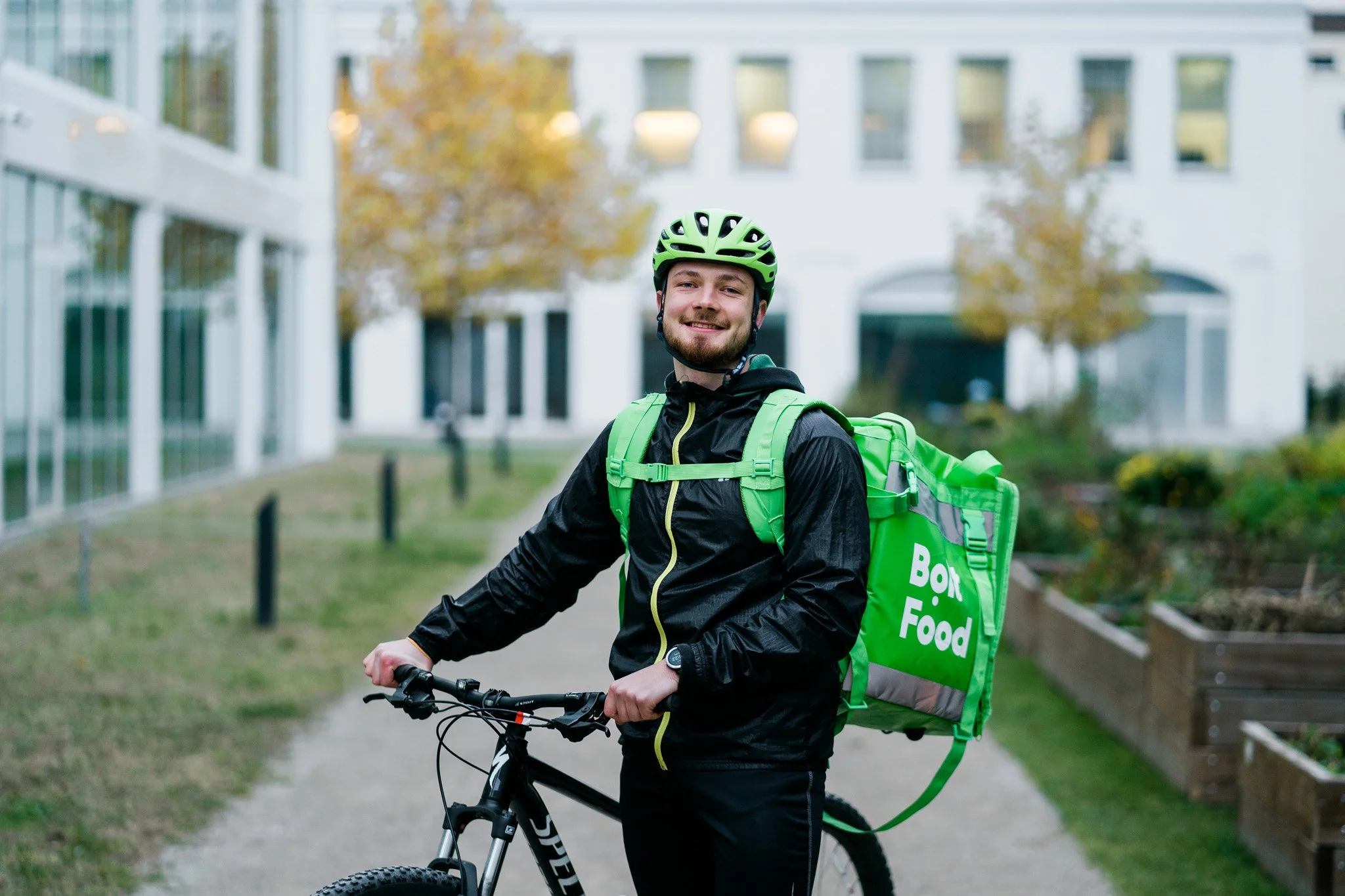 A smiling young man in a black jacket and green helmet standing on a mountain bike, carrying a large green food delivery bag labeled 'Box Food,' outdoors near modern buildings and trees with autumn foliage. Advertising campaign photographer Zurich.