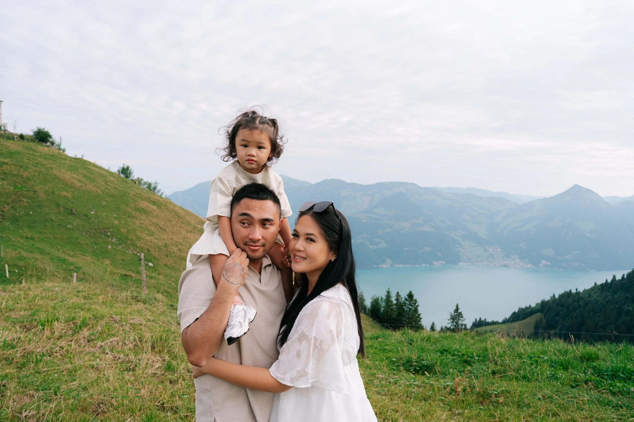 A family of three enjoying nature on a hillside with a lake and mountains in the background. The father carries a young girl on his shoulders, while the mother smiles beside him. Family photoshoot in Switzerland.