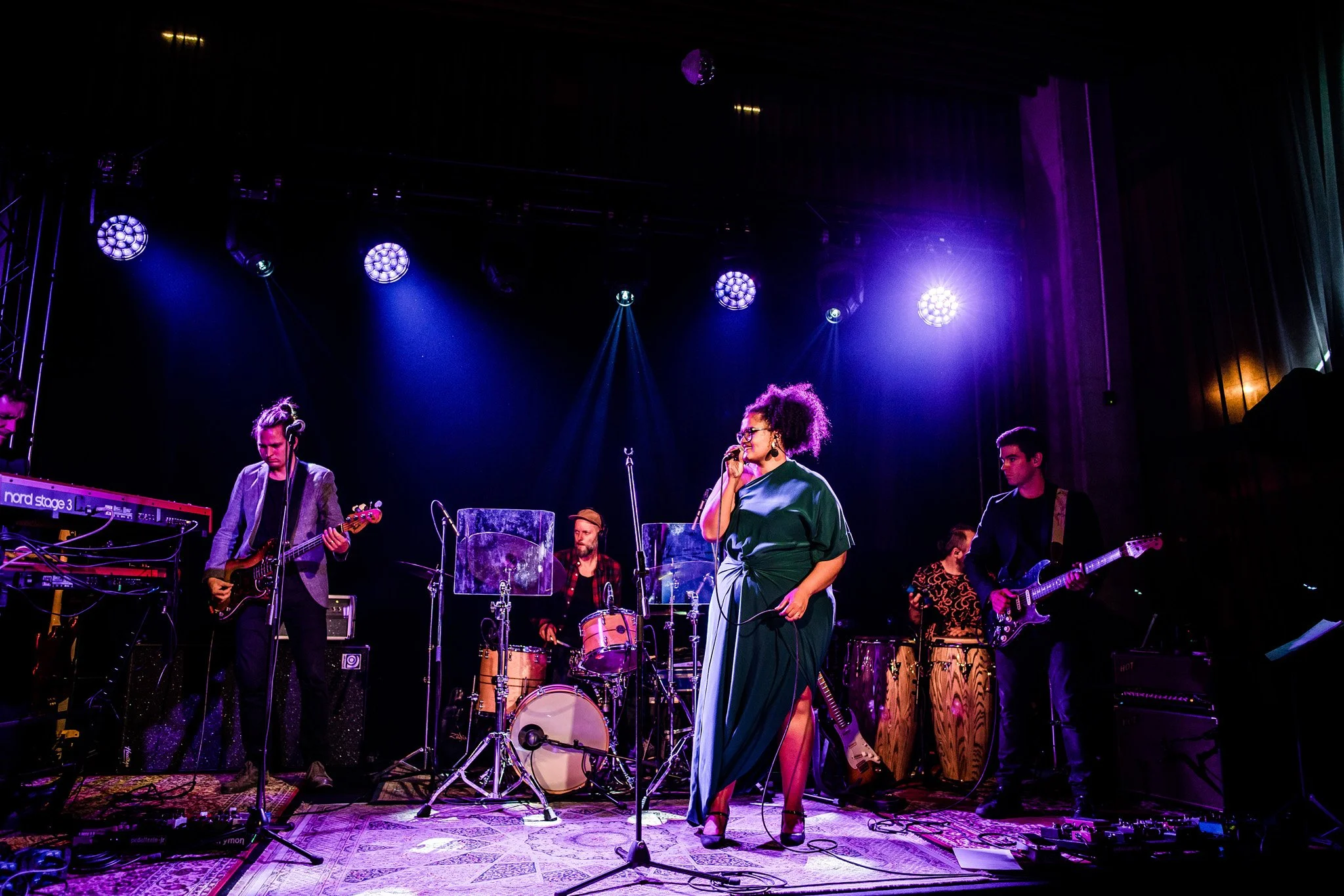A band performs on stage with purple and blue lighting, featuring a singer in a green dress, guitarists, a drummer, and percussionists. Concert and singer photography in Zürich, Zug, Bern, Lucerne, Basel, St. Gallen and across Switzerland.