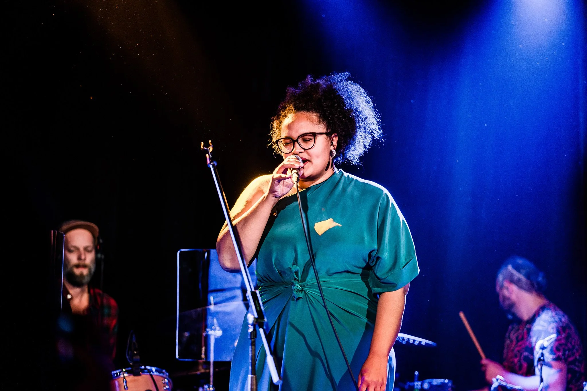 A woman with curly hair, glasses, and earrings singing into a microphone on stage, with two band members playing instruments in the background. Concert photography in Zürich, Zug, Bern, Lucerne, Basel, St. Gallen and across Switzerland.