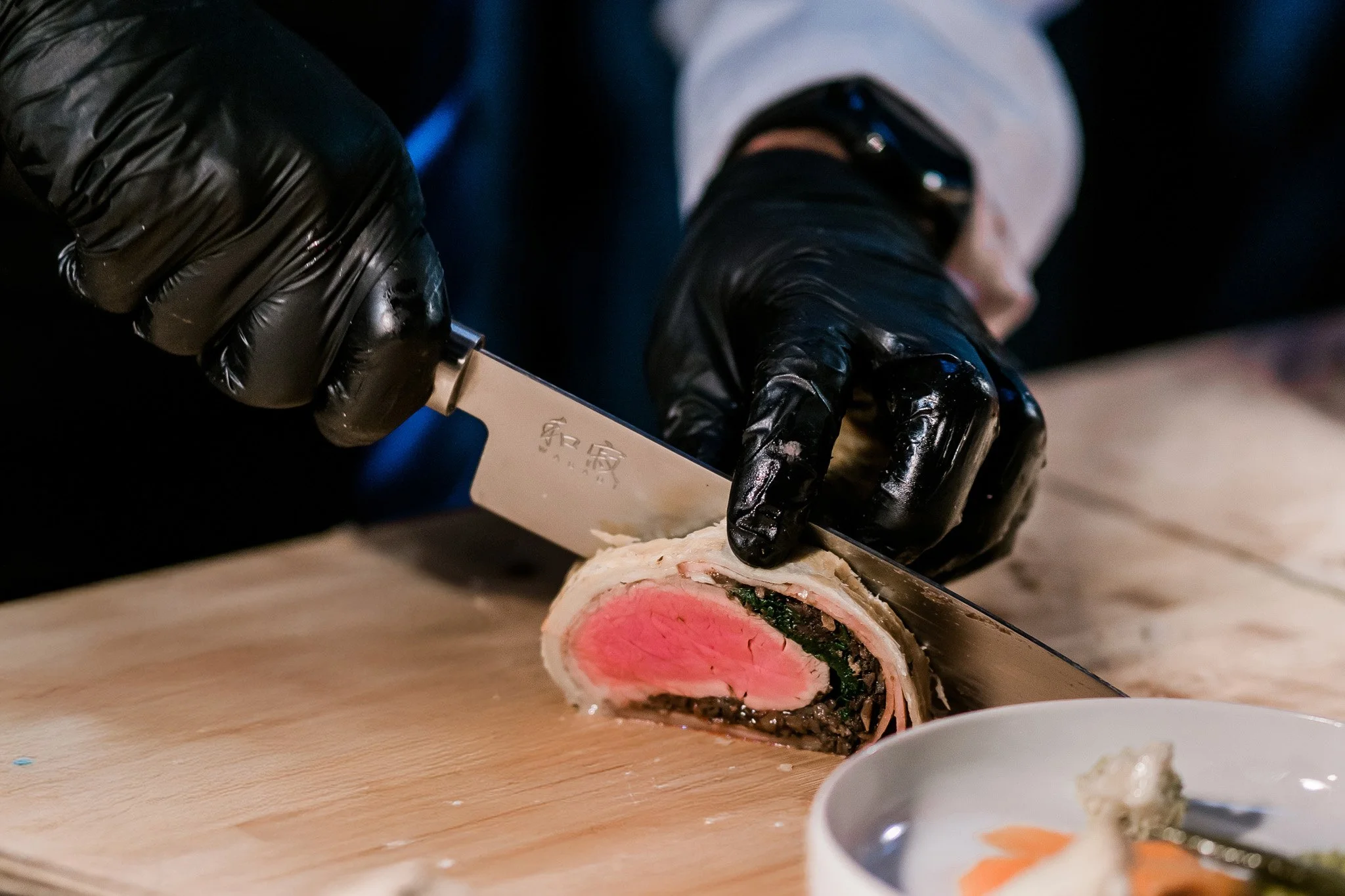 A person wearing black gloves is slicing a piece of beef Wellington on a wooden cutting board. Catering and food photography in Zürich, Zug, Bern, Lucerne, Basel, St. Gallen and across Switzerland.