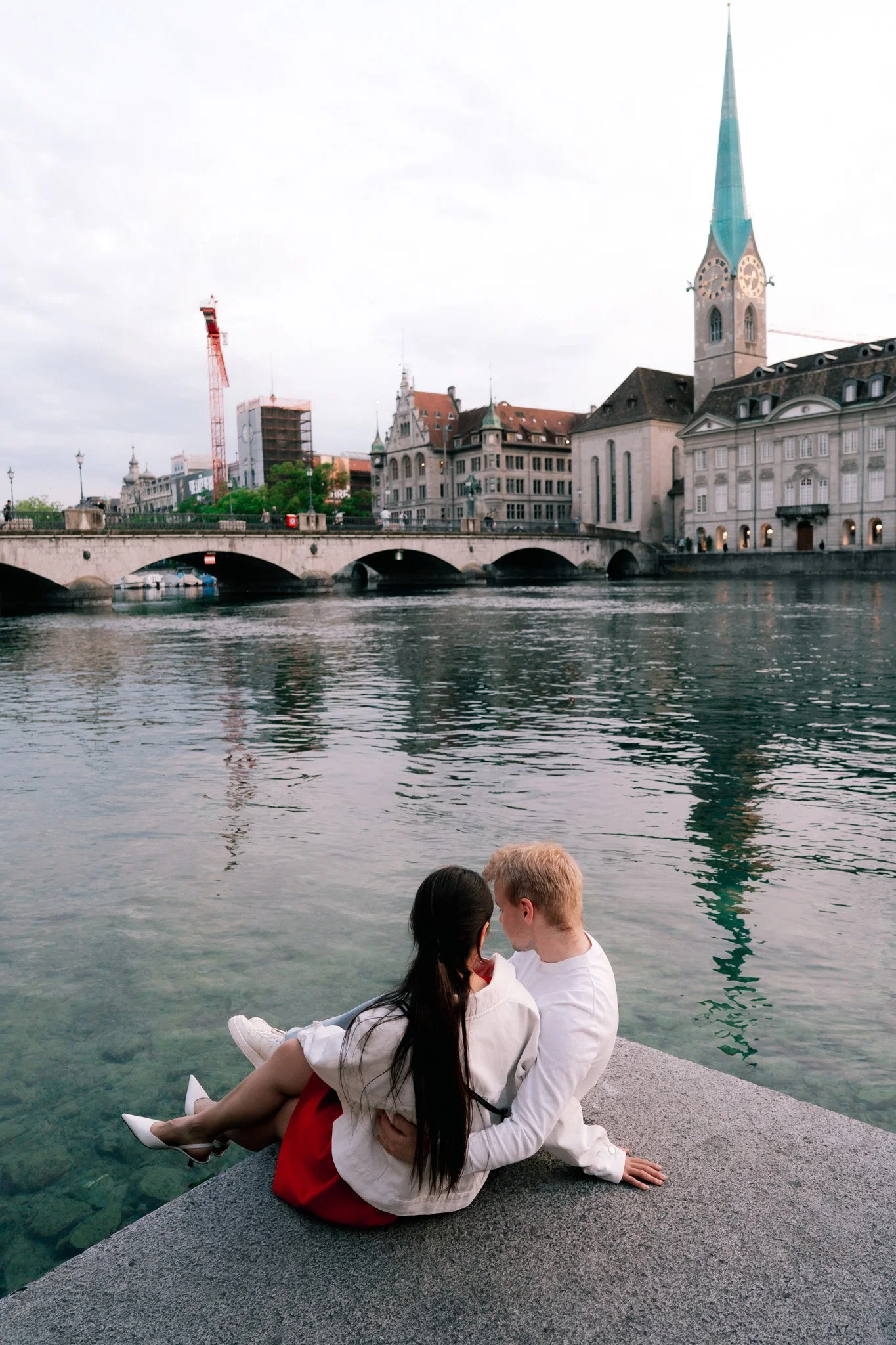 A couple sitting by a river with historic buildings in Zurich Switzerland and a church in the background.
