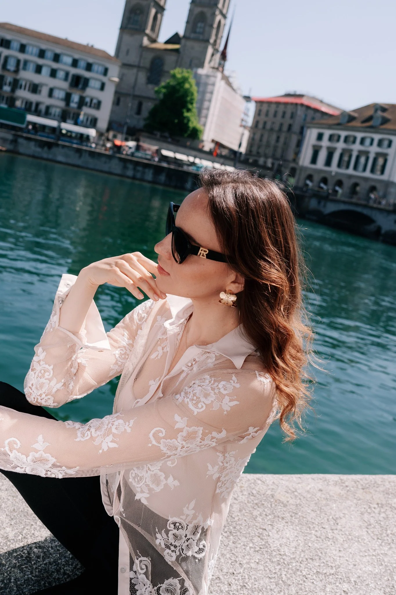 A woman with wavy brown hair wearing large black sunglasses, floral earrings, and a sheer white lace top sitting by a body of water with historic buildings and a church in the background on a sunny day.