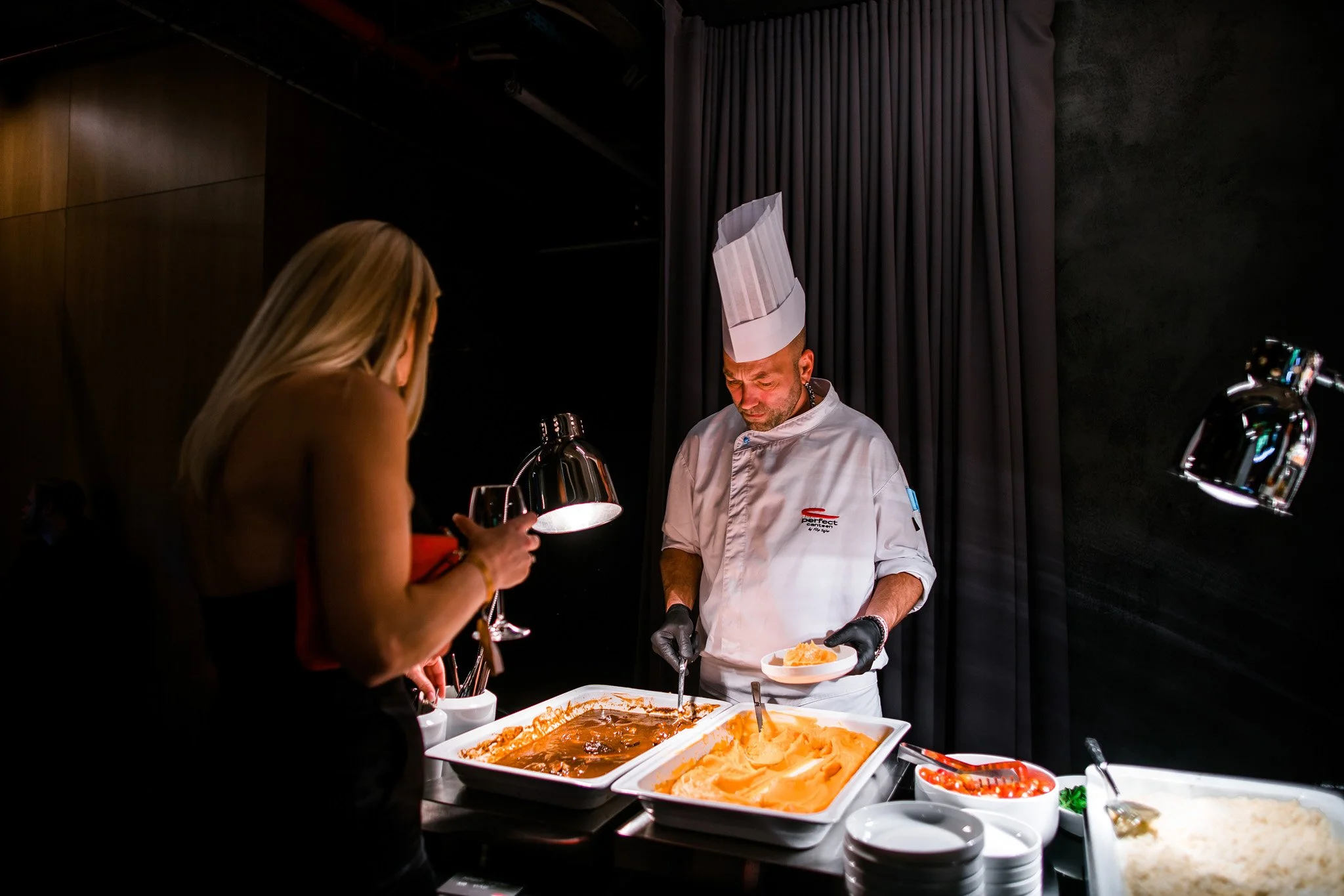 Chef serving food from large trays to a woman at a buffet-style setting with dark background and curtains. Catering, culinary and food photography in Zürich, Zug, Bern, Lucerne, Basel, St. Gallen and across Switzerland.