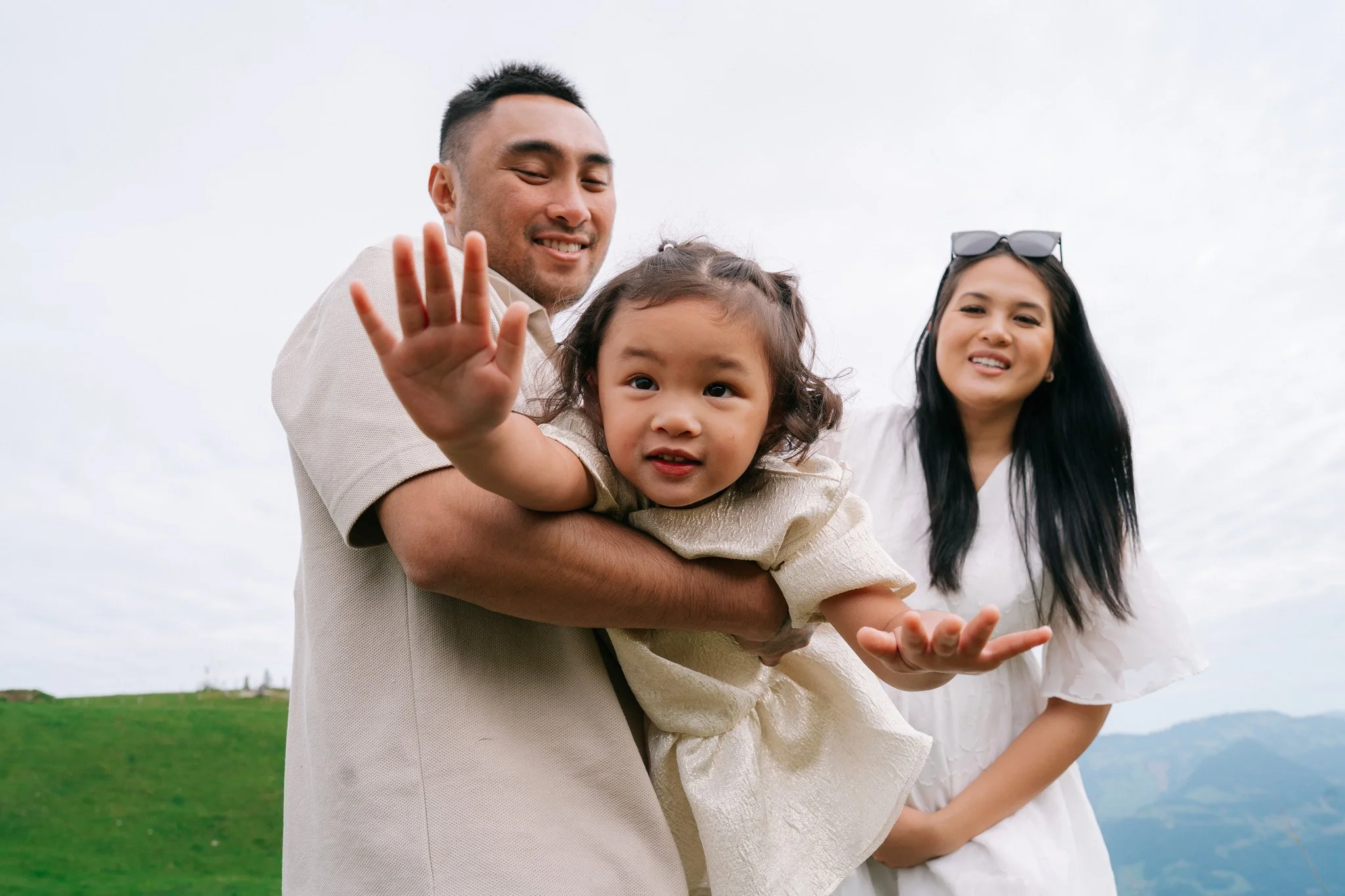 Little girl being held up by a man, waving at the camera, with woman smiling nearby outside on a cloudy day. Family photoshoot in Switzerland.