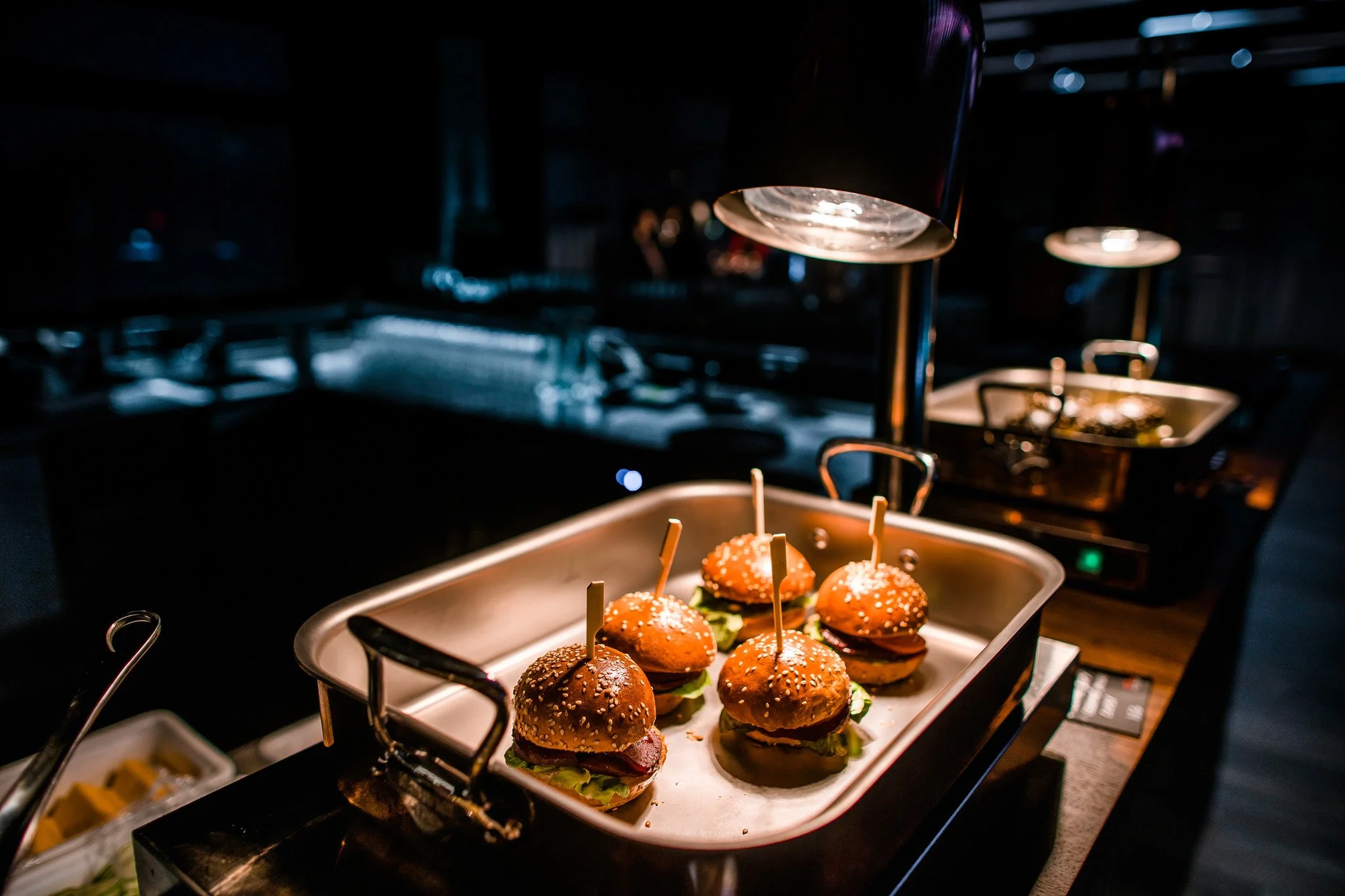 Six mini cheeseburgers with lettuce, bacon, and sesame seed buns on a stainless steel tray in a dimly lit buffet setting. Catering and food photography in Zürich, Zug, Bern, Lucerne, Basel, St. Gallen and across Switzerland.