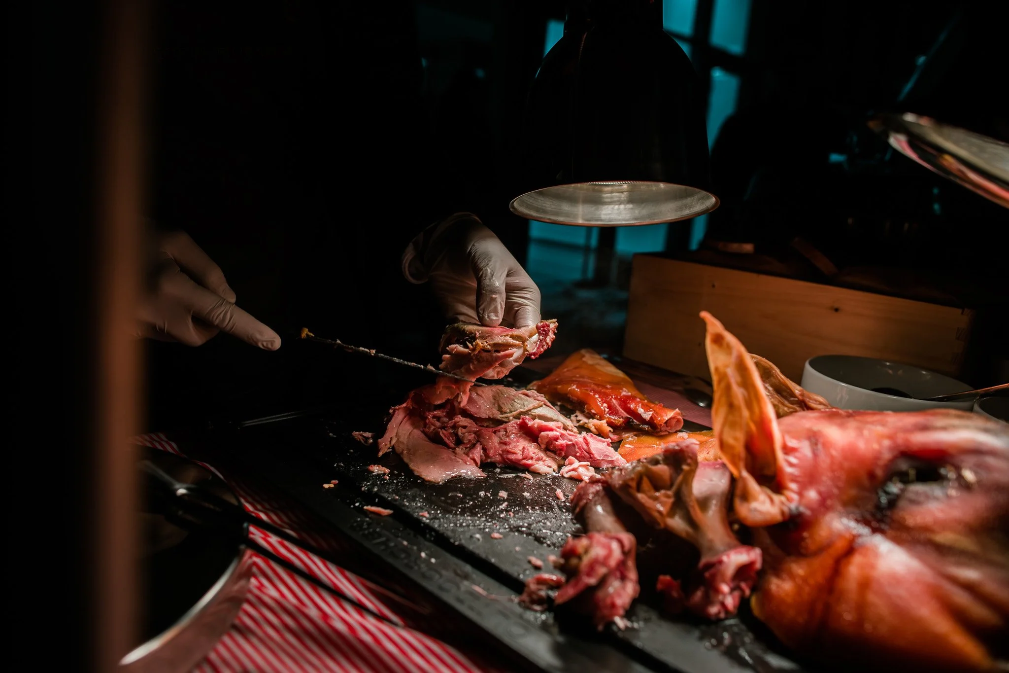 Cook wearing gloves carving a large piece of meat on a black cutting surface, with other meat pieces nearby, in a dimly lit kitchen or butcher shop. Catering and food photography in Zürich, Zug, Bern, Lucerne, Basel, St. Gallen and across Switzerland