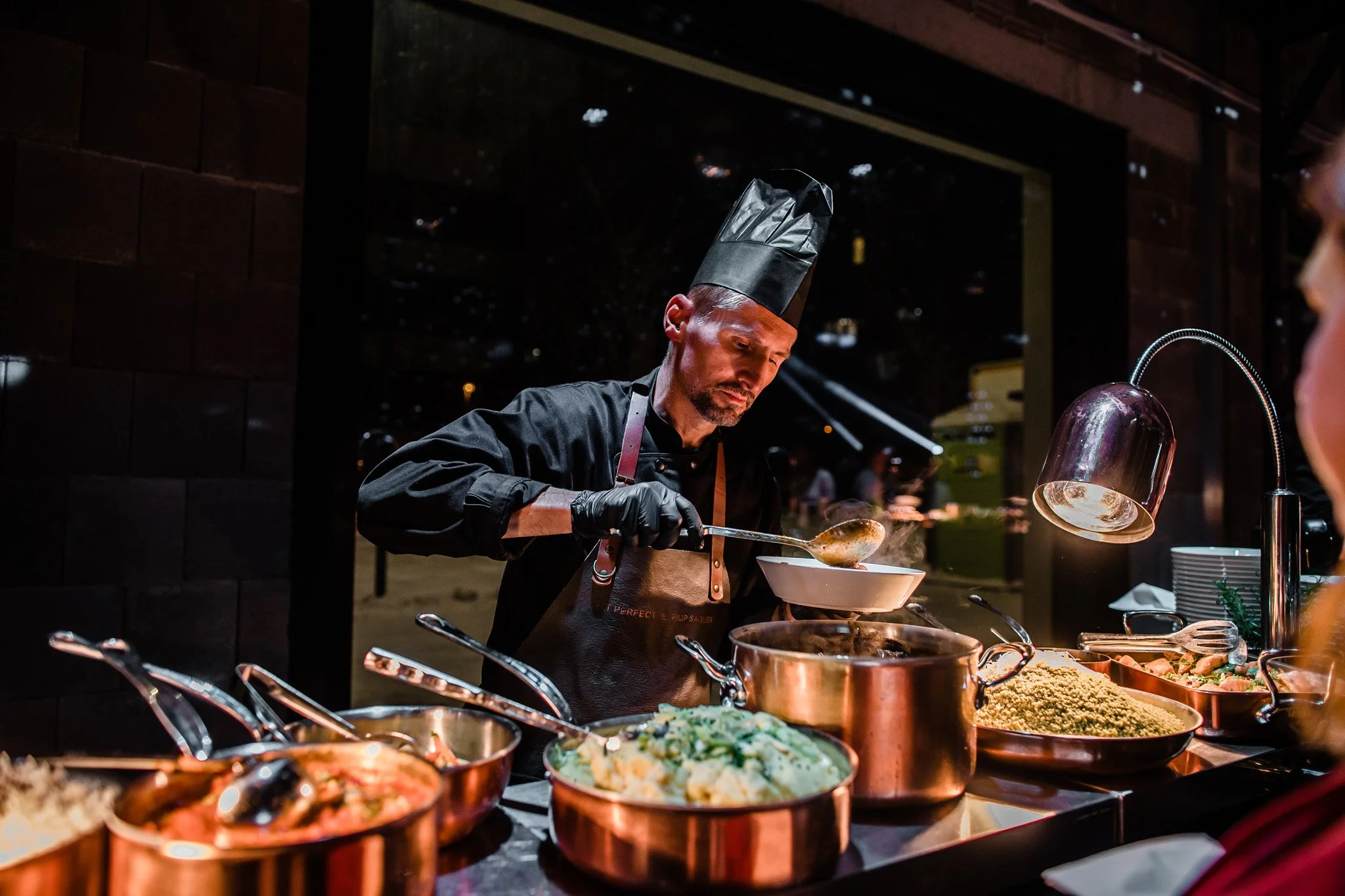 A chef in a black uniform and tall black hat plates food at a buffet station with several copper pots and bowls filled with various dishes. The chef is using a ladle to serve food into a bowl, with a warm glow from a silver heat lamp overhead. 
