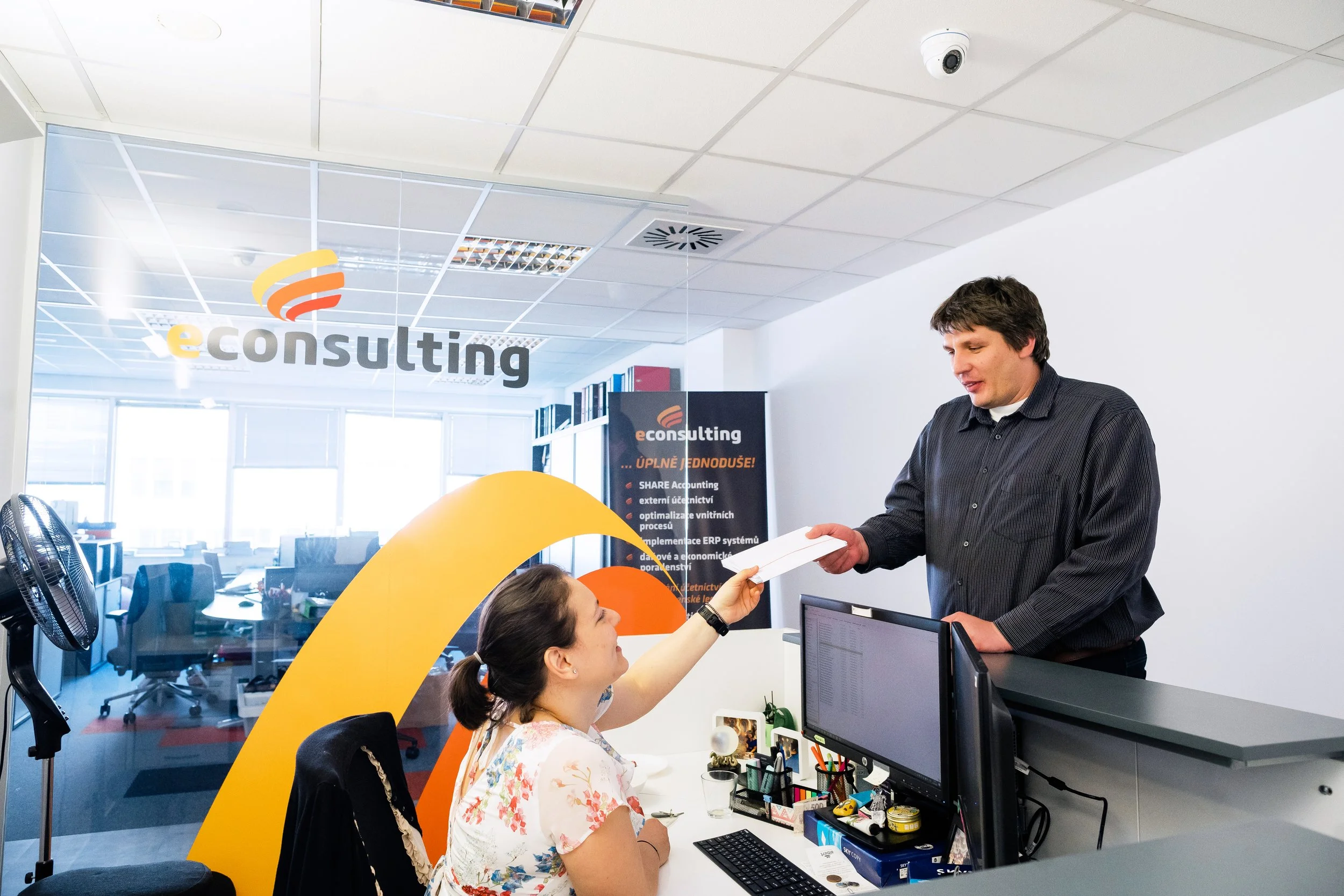 A woman at a reception desk is reaching out to take a white box from a man standing on the other side of the desk in an office environment. The office has a glass wall with the logo and a colorful banner with company information, computer monitors, a