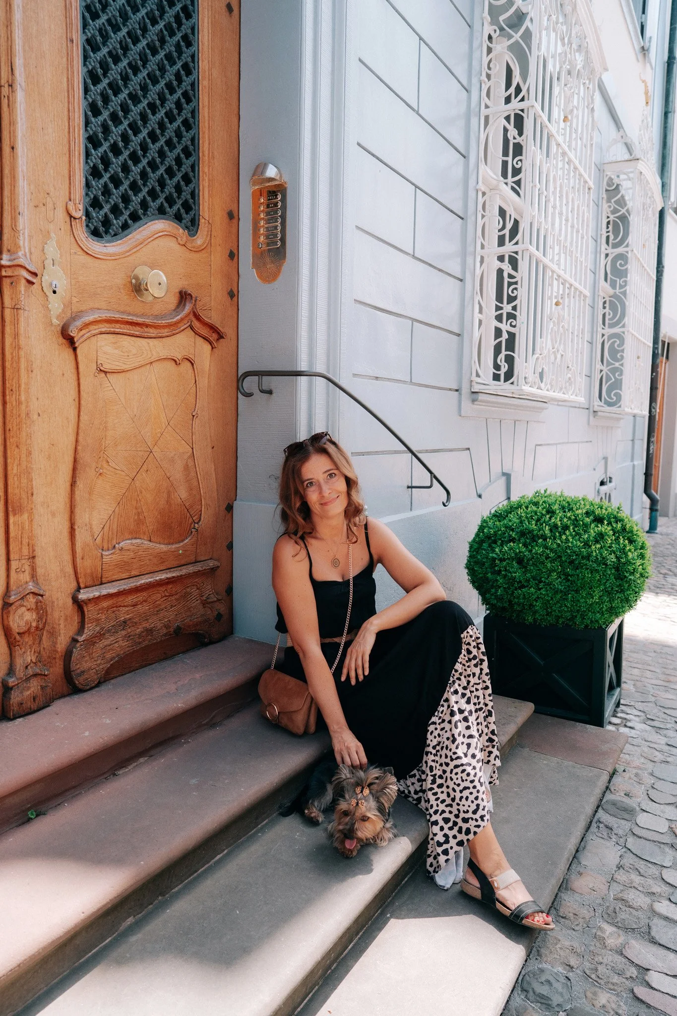 Lifestyle photoshoot of a woman sitting on stone steps outside a building in Basel, Switzerland, with a small dog at her feet, colorful street in the background, and a large green plant nearby.