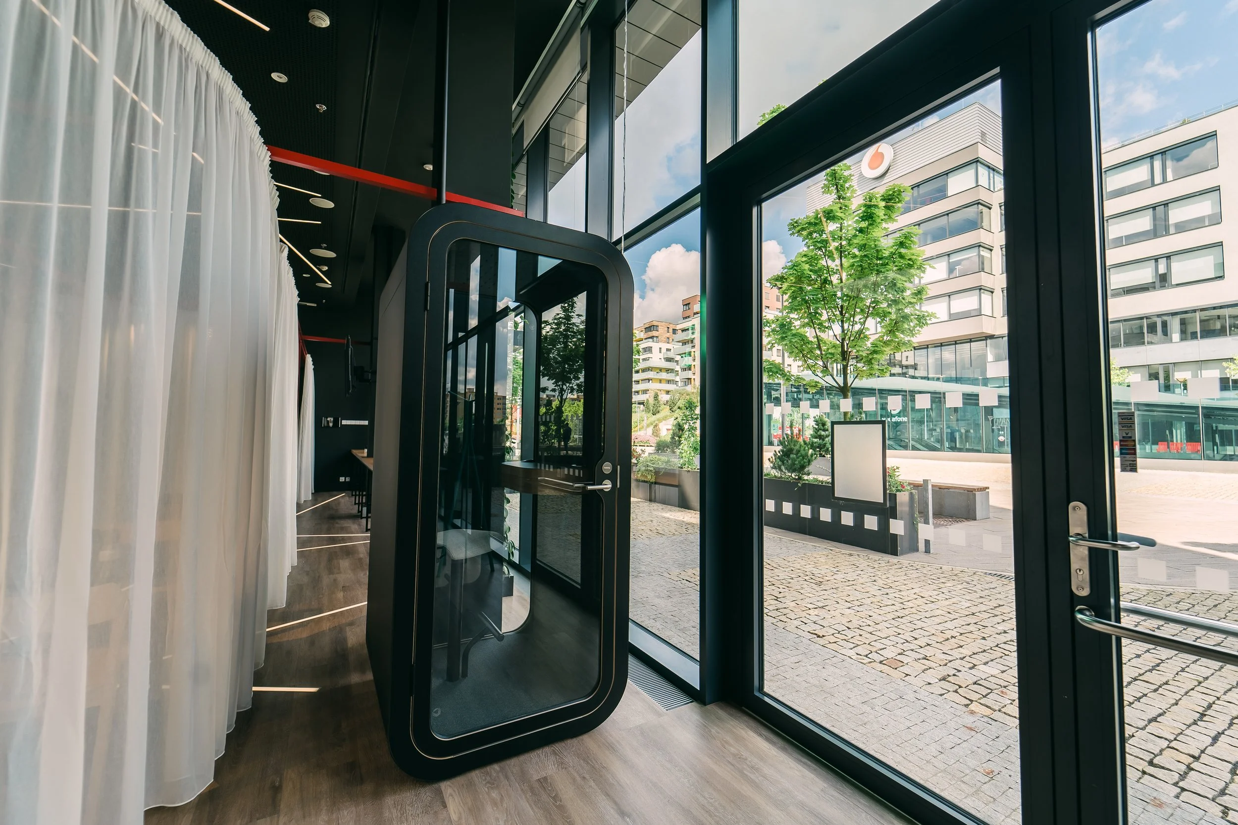 Modern interior with a glass entrance door leading outside to a street with trees and buildings. White curtains on the left side, and a photo booth or privacy pod near the door. Restaurant interior photography in Switzerland.
