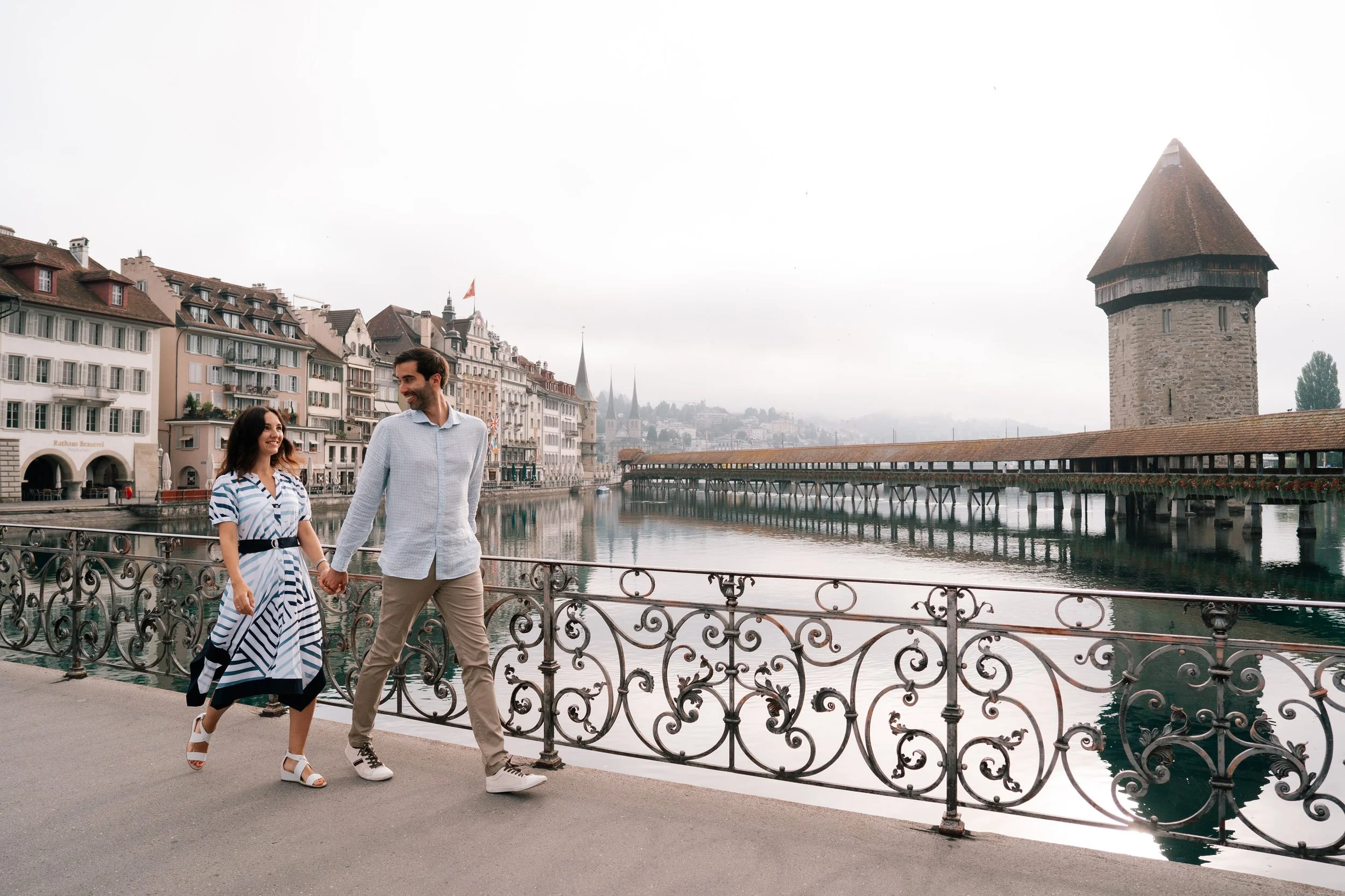 A smiling couple walking hand-in-hand along a riverside promenade in Lucerne, Switzerland, with historic buildings and the Water Tower visible in the background.