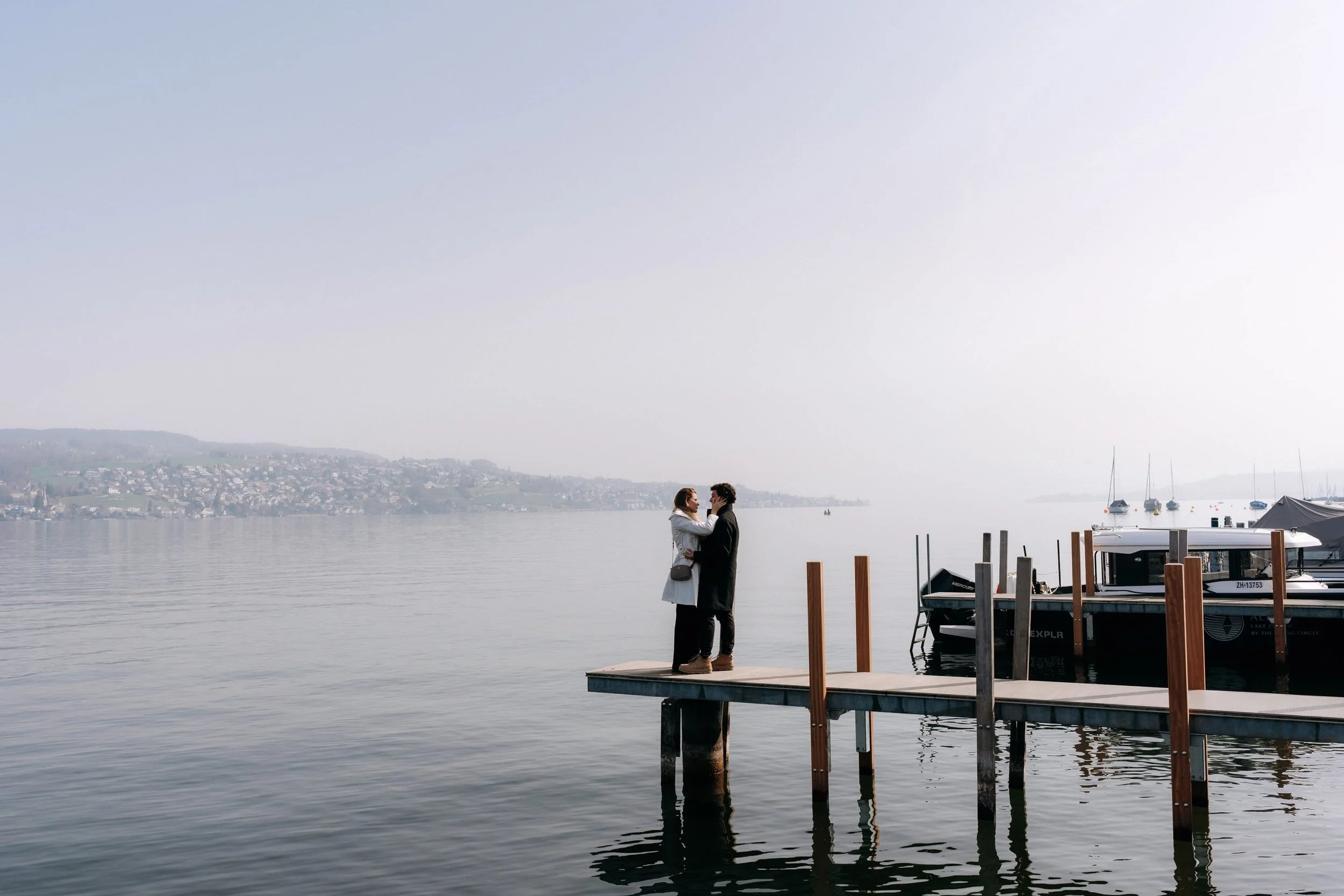 A couple standing on a dock by the water near Zurichsee, after an engagement photoshoot.