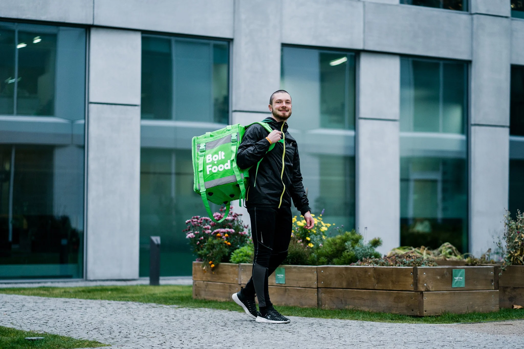 A man with a beard dressed in black athletic clothing carrying a green Bolt Food delivery backpack walking on a paved sidewalk with a modern building. Advertising campaign photographer for corporate and startup business Switzerland.