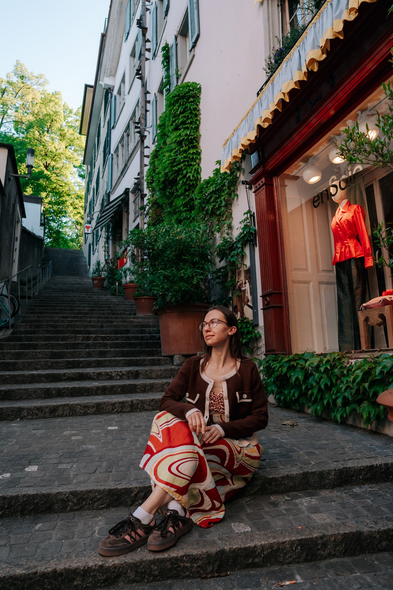 A woman sitting on outdoor stairs in historical city centre of Zurich, Switzerland, with shops and greenery around her doing a vacation photoshoot.