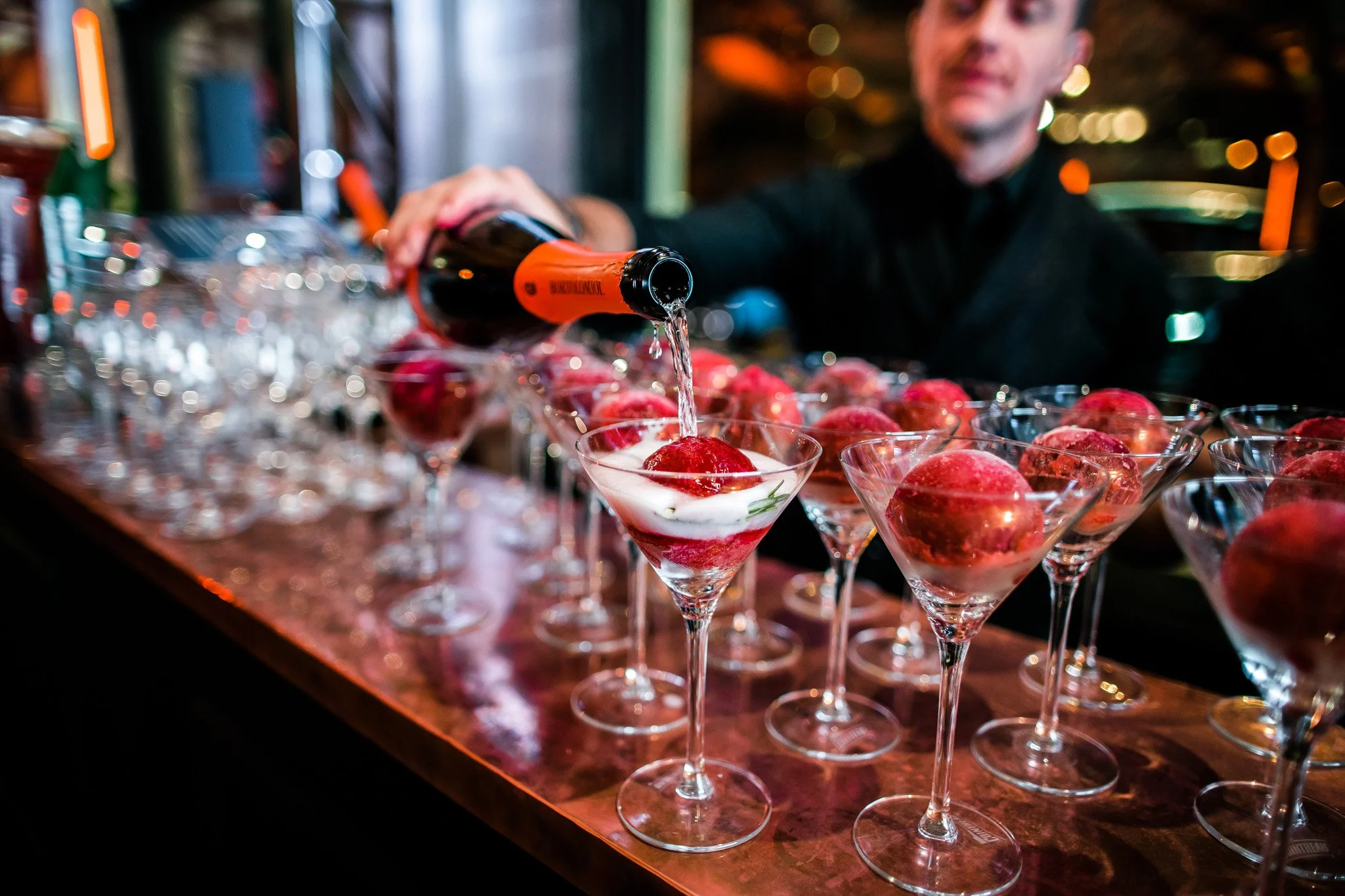A bartender pouring champagne into a martini glass with raspberries and cream on a bar counter filled with similar glasses, in a dimly lit setting. Catering and food photography in Zürich, Zug, Bern, Lucerne, Basel, St. Gallen and across Switzerland.