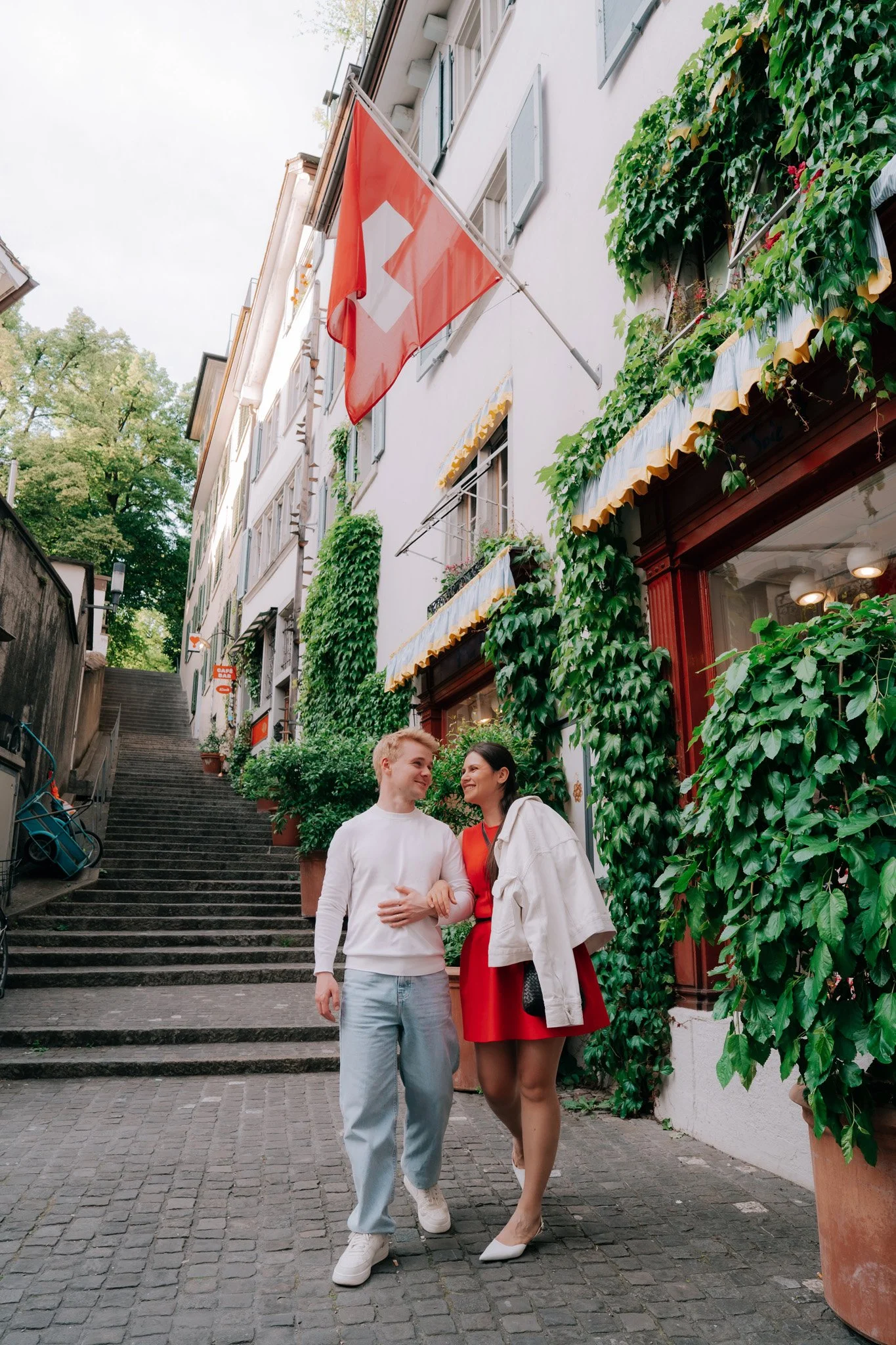 A couple love story, walking togehter in arm down a cobblestone street in a European city, with a Swiss flag hanging above them and lush greenery surrounding the buildings.