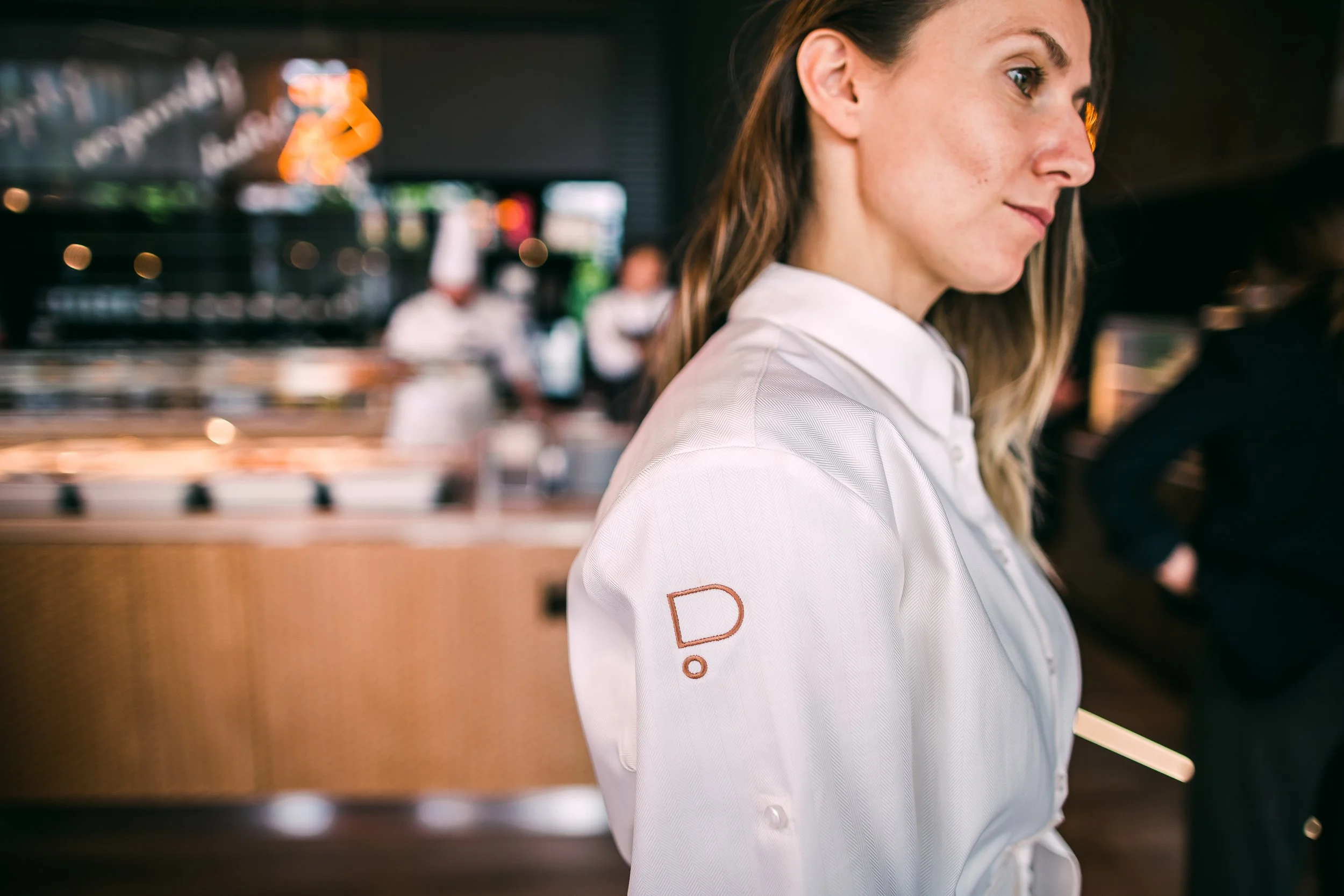 Close-up of a female restaurant employee wearing a white shirt with a logo on the sleeve, working in a restaurant with blurred background of chefs and kitchen. Restaurant interior photography in Zürich, Zug, Bern, Lucerne, Basel, Switzerland.