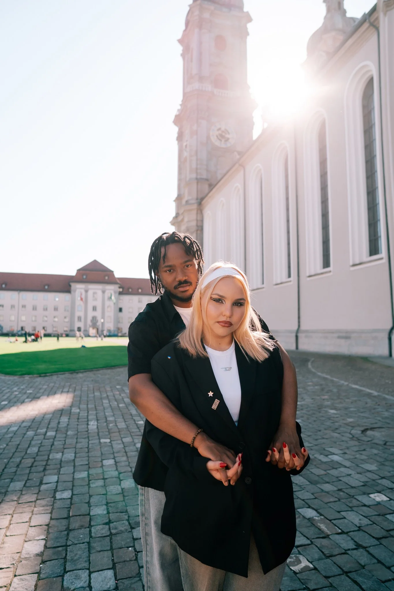 A man and woman have romantic photoshoot in cobblestone pathway in front of a historic building in St Gallen Switzerland with a clock tower, with the sunlight shining from behind them.