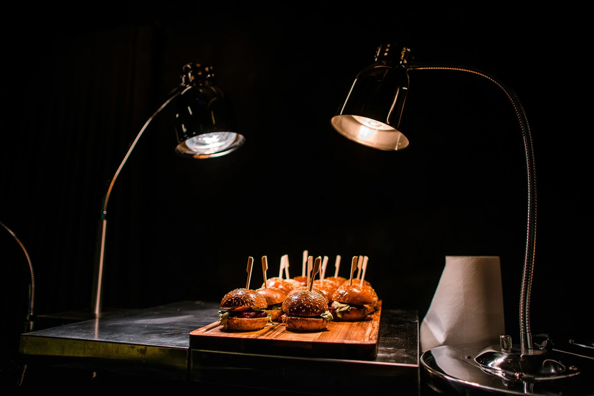 A wooden cutting board with multiple mini burgers topped with toothpicks, illuminated by two adjustable desk lamps in a dark setting. Catering, culinary and food photography in Zürich, Zug, Bern, Lucerne, Basel, St. Gallen and across Switzerland.