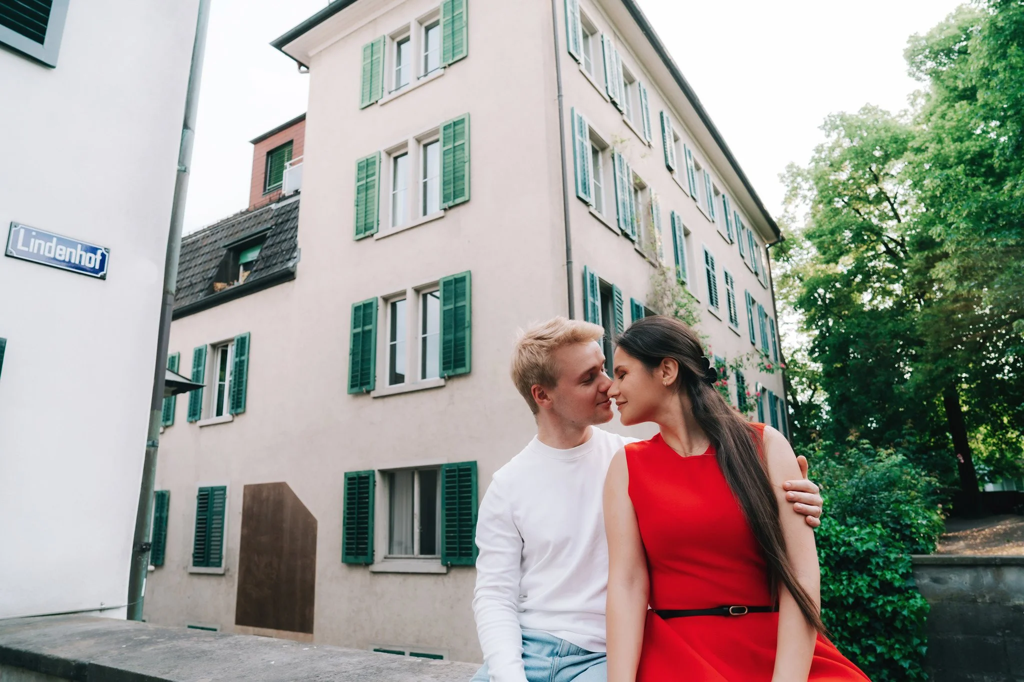A young couple sitting close together outdoors in Zurich historic centre near a building with green window shutters, leaning in for a gentle, face-to-face moment, with trees in the background.