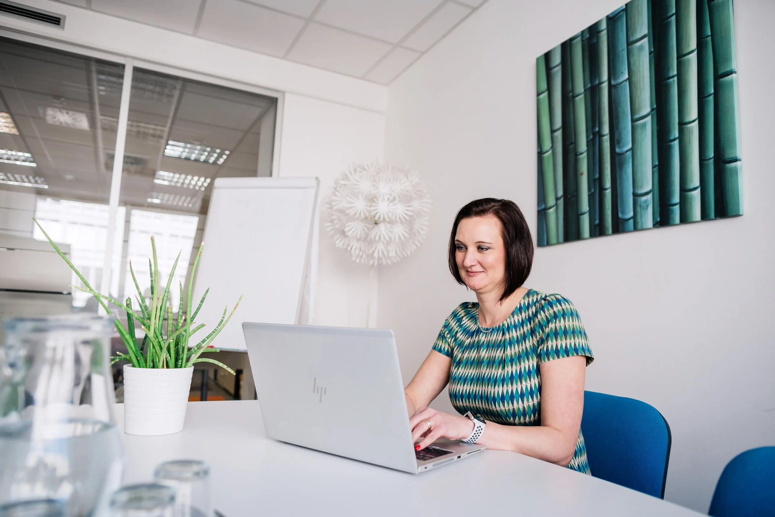 A woman with short dark hair and a patterned dress sitting at a conference table, working on a silver laptop, in a modern office with a white wall, green framed artwork, a white leafy decor, a potted plant, glasses and a mirror in the background.