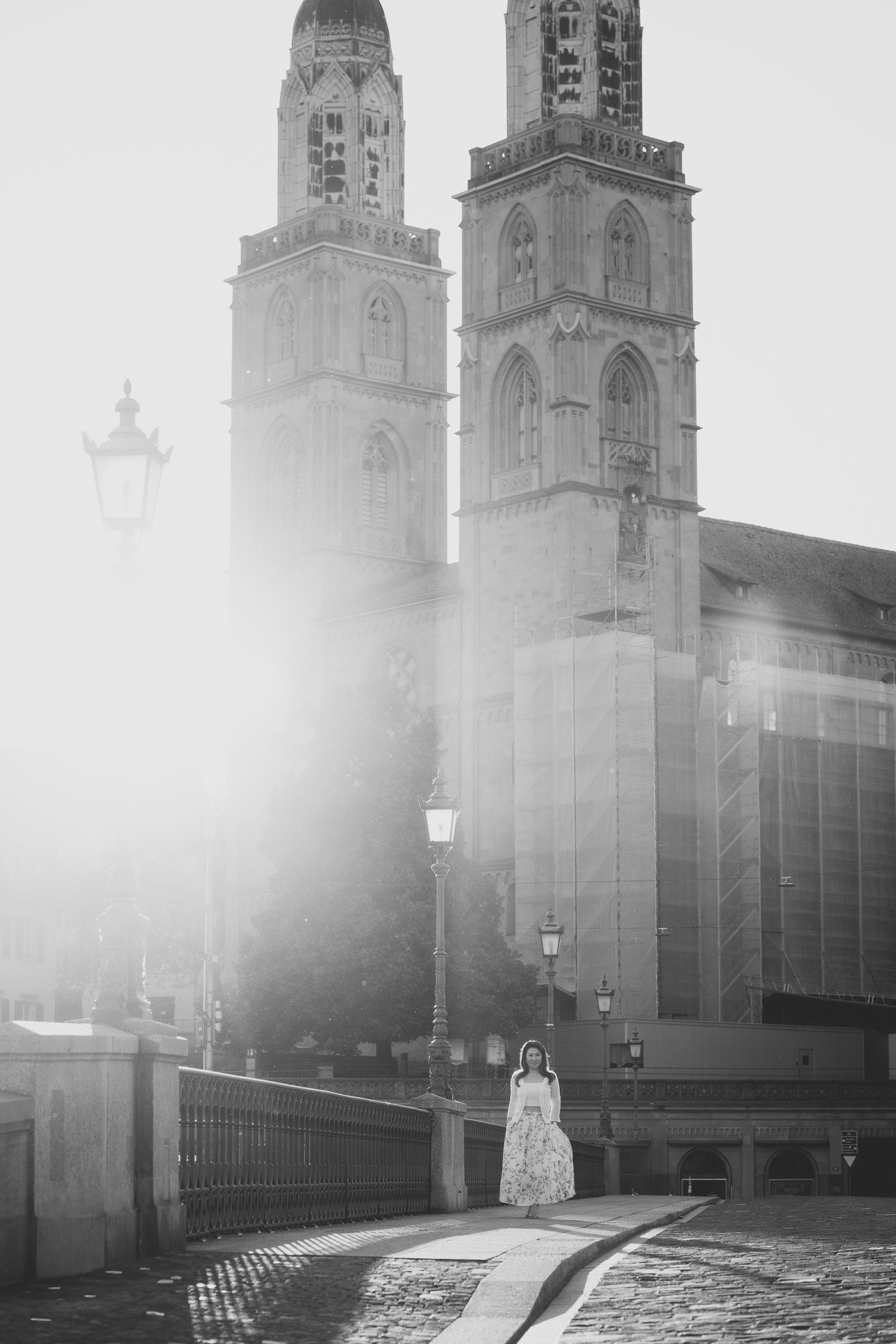 Black and white photo of a woman standing on a cobblestone street with tall church towers in the background, bright sunlight creating lens flare.