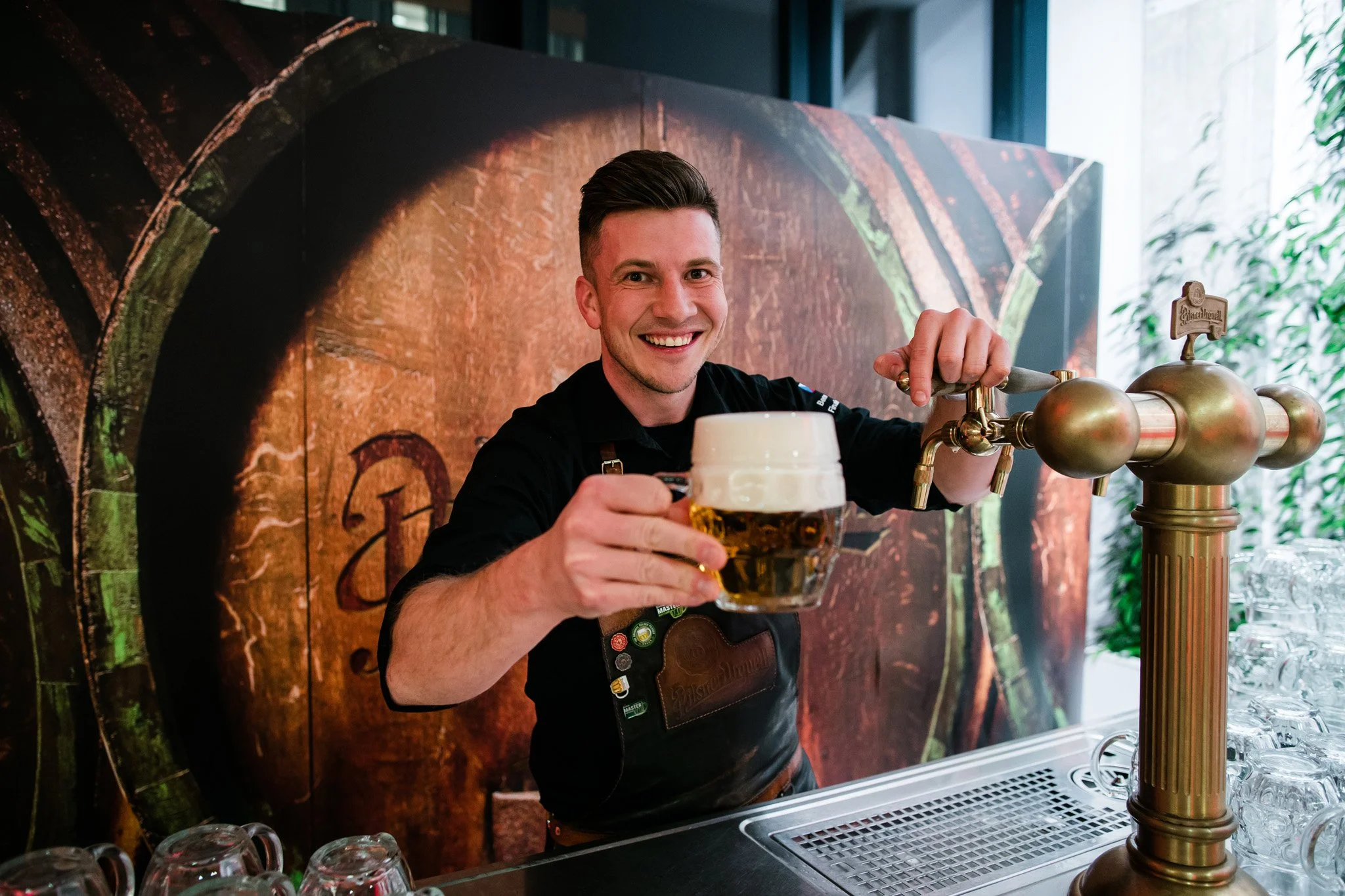 A smiling bartender pours a pint of beer from a tap at a bar with a copper-colored background. Catering and food photography in Zürich, Zug, Bern, Lucerne, Basel, St. Gallen and across Switzerland.