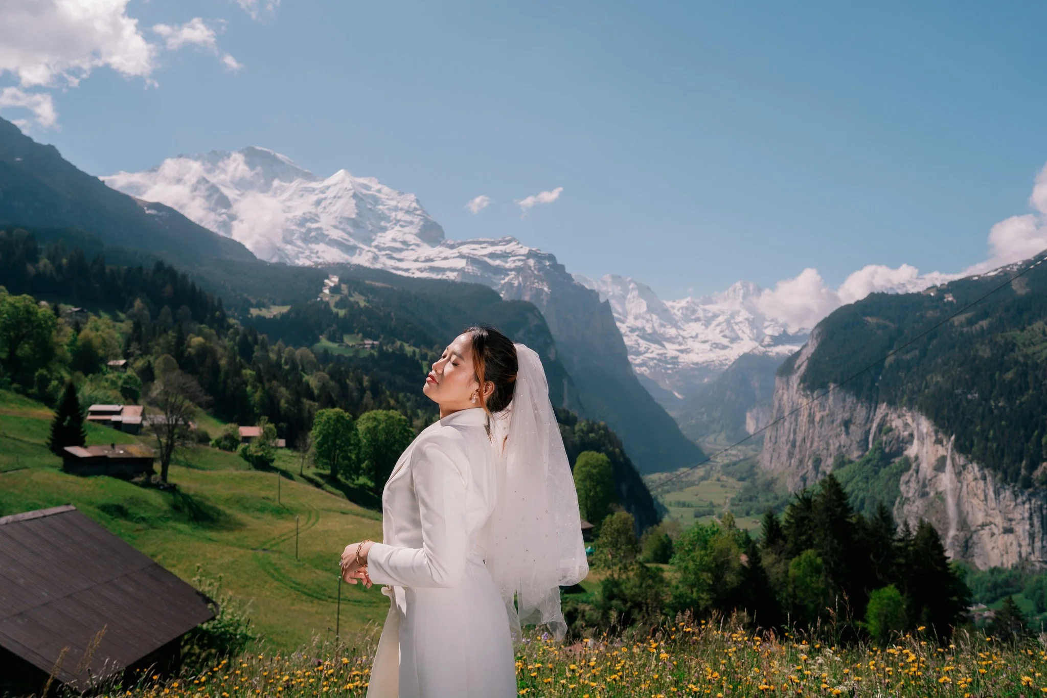 Woman doing wedding photoshoot standing on Swiss grassy hillside of Wengen, Lauterbrunnen, Switzerland with yellow flowers, mountains and snow-capped peaks in the background.