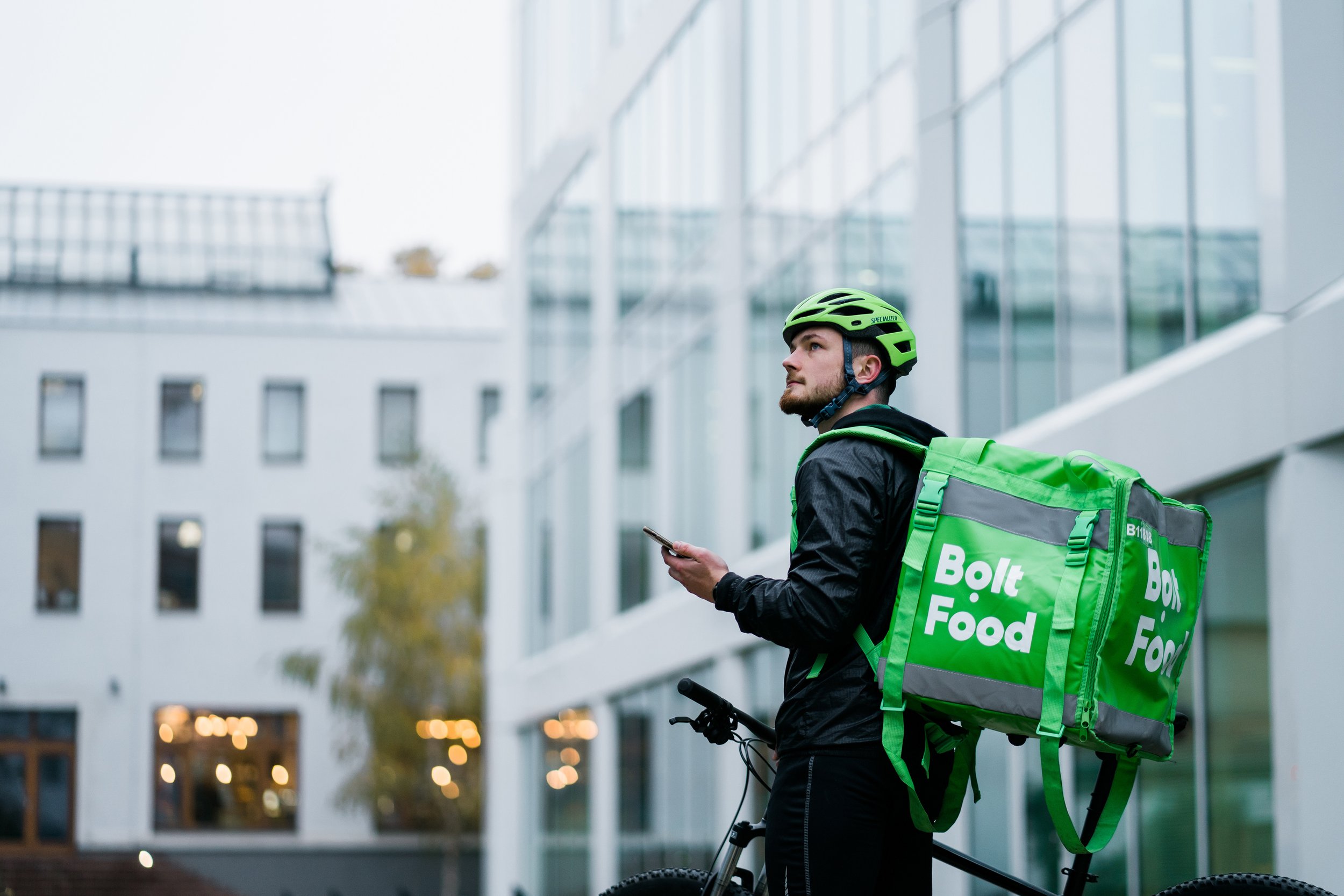 A man in a black jacket with a green helmet and a large green Bolt Food delivery backpack, standing next to a bicycle, looking at his phone outside a modern building. Advertising photography in Zürich, Zug, Lucerne, St. Gallen and across Switzerland.